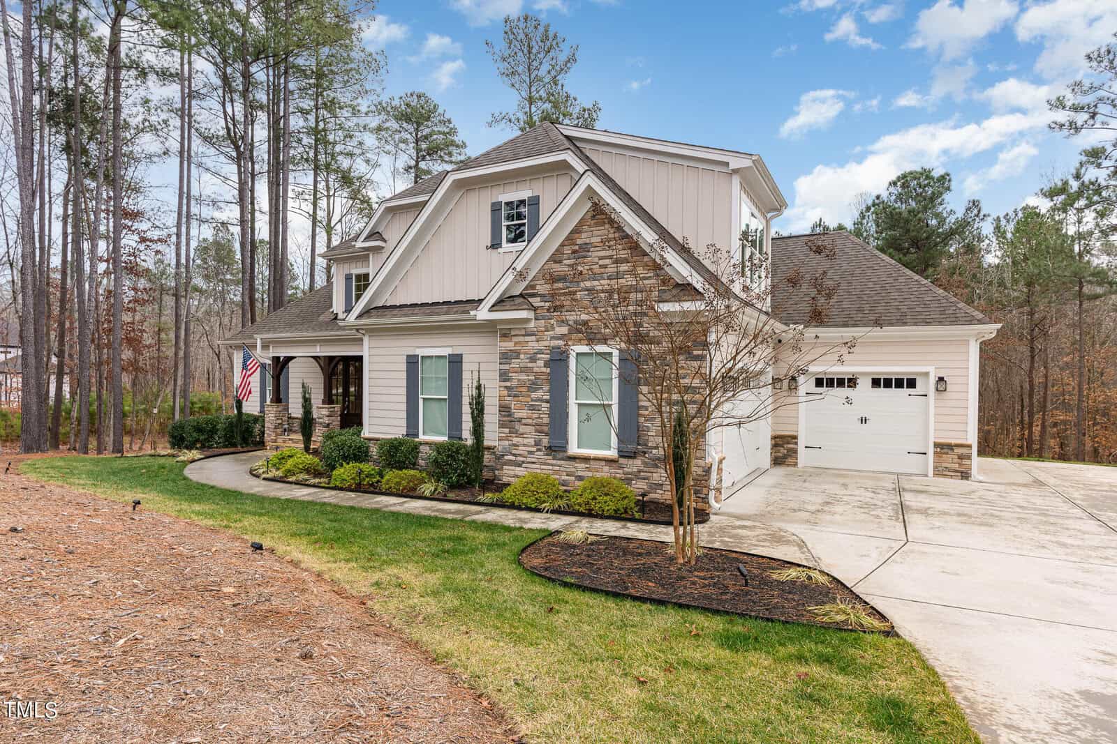 A two-story house with stone and siding exterior, featuring a garage and front porch. The home is surrounded by tall trees and a well-maintained lawn. An American flag hangs near the porch, and the sky is partly cloudy.