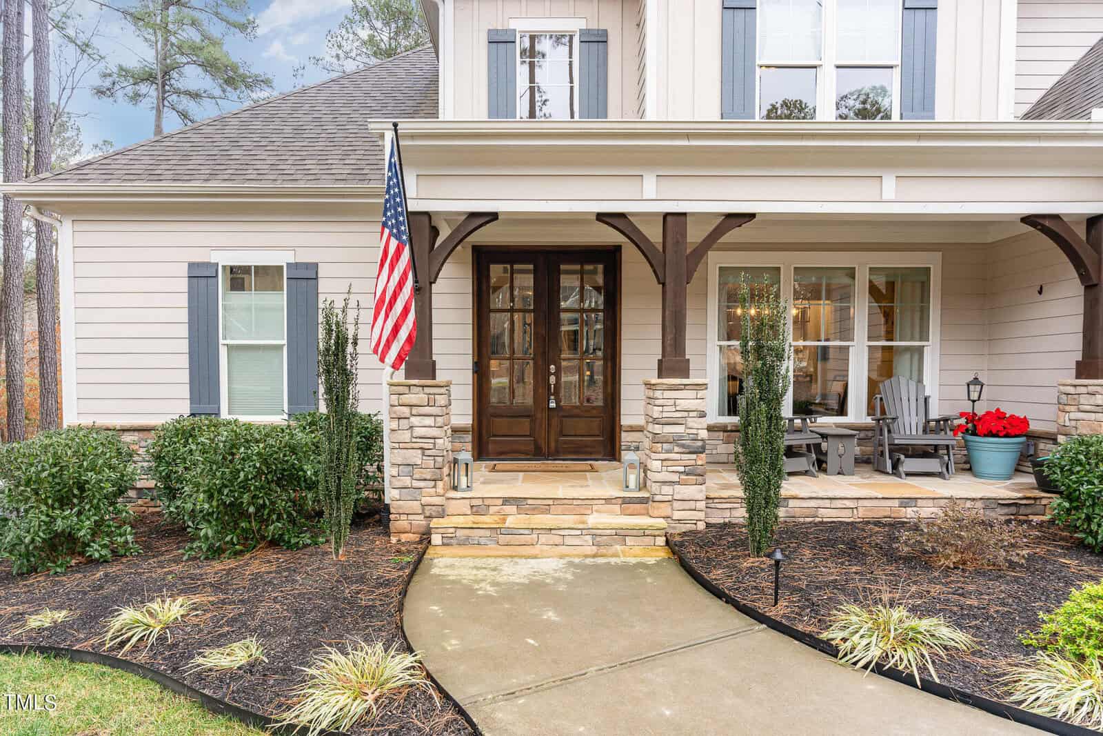 A charming house with a stone porch, wooden beams, and an American flag. Gray siding and white trim complement the windows, flanked by dark shutters. The front yard is landscaped with shrubs and a walkway leading to the entrance.