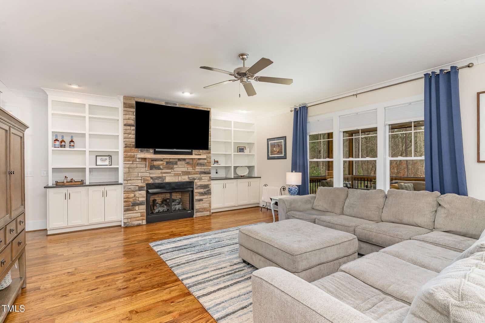 A cozy living room with a large beige sectional sofa, a striped rug, and a stone fireplace topped with a TV. Built-in shelves frame the fireplace, and large windows with blue curtains let in natural light. A ceiling fan is overhead.