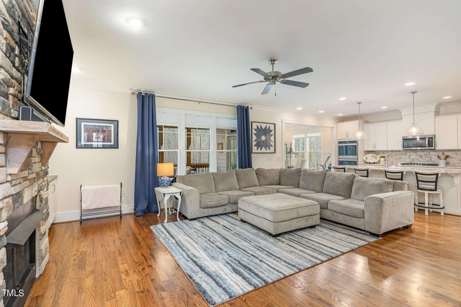 Spacious living room with a gray sectional sofa on a blue-striped rug, a wall-mounted TV, and a stone fireplace. The room features wooden floors, a ceiling fan, large windows with blue curtains, and an open view of a modern kitchen with barstools.