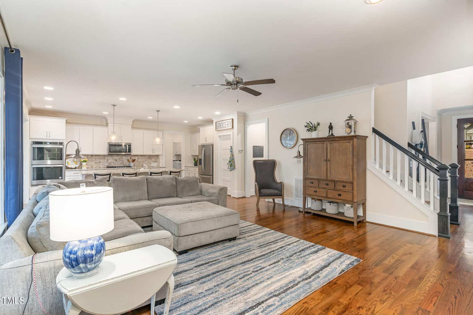 Spacious living room with a large gray sectional sofa, blue and white rug, and a ceiling fan. Open concept kitchen in the background features white cabinets and a central island with stools. A wooden side cabinet is near the staircase.