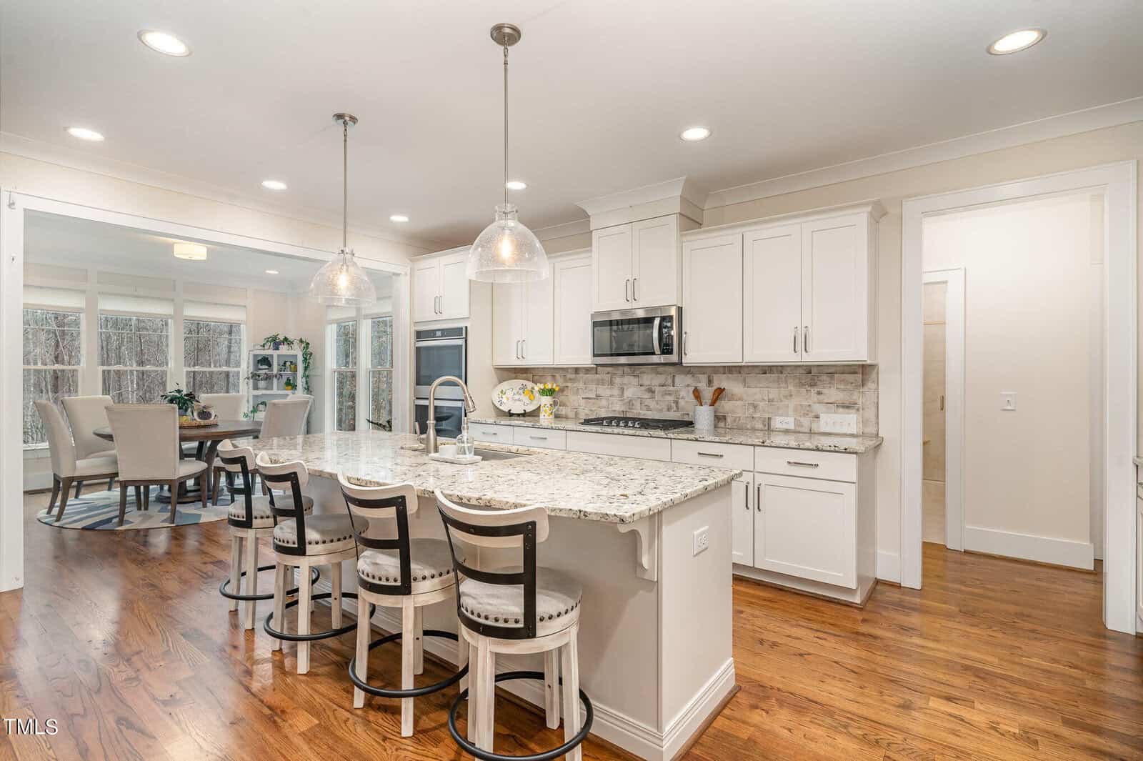 A modern kitchen with white cabinets and granite countertops. It features a large island with four cushioned barstools, stainless steel appliances, and pendant lighting. A dining area with a table and chairs is visible in the background.