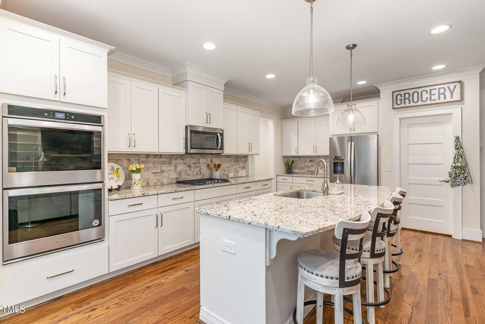 A bright, modern kitchen with white cabinets, stainless steel appliances, and granite countertops. There's an island with four cushioned chairs and pendant lights above. A "Grocery" sign hangs on the wall near a closed door.