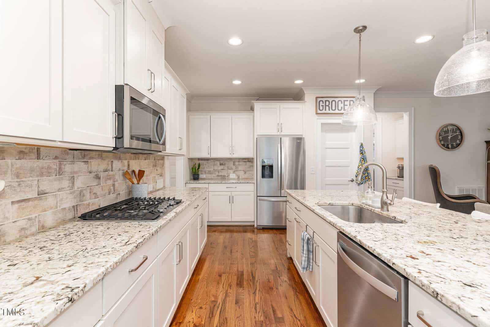 A modern kitchen featuring white cabinets, stainless steel appliances, granite countertops, and a center island with a sink. The room is well-lit with pendant lights and has hardwood flooring and a "GROCERY" sign on the wall.