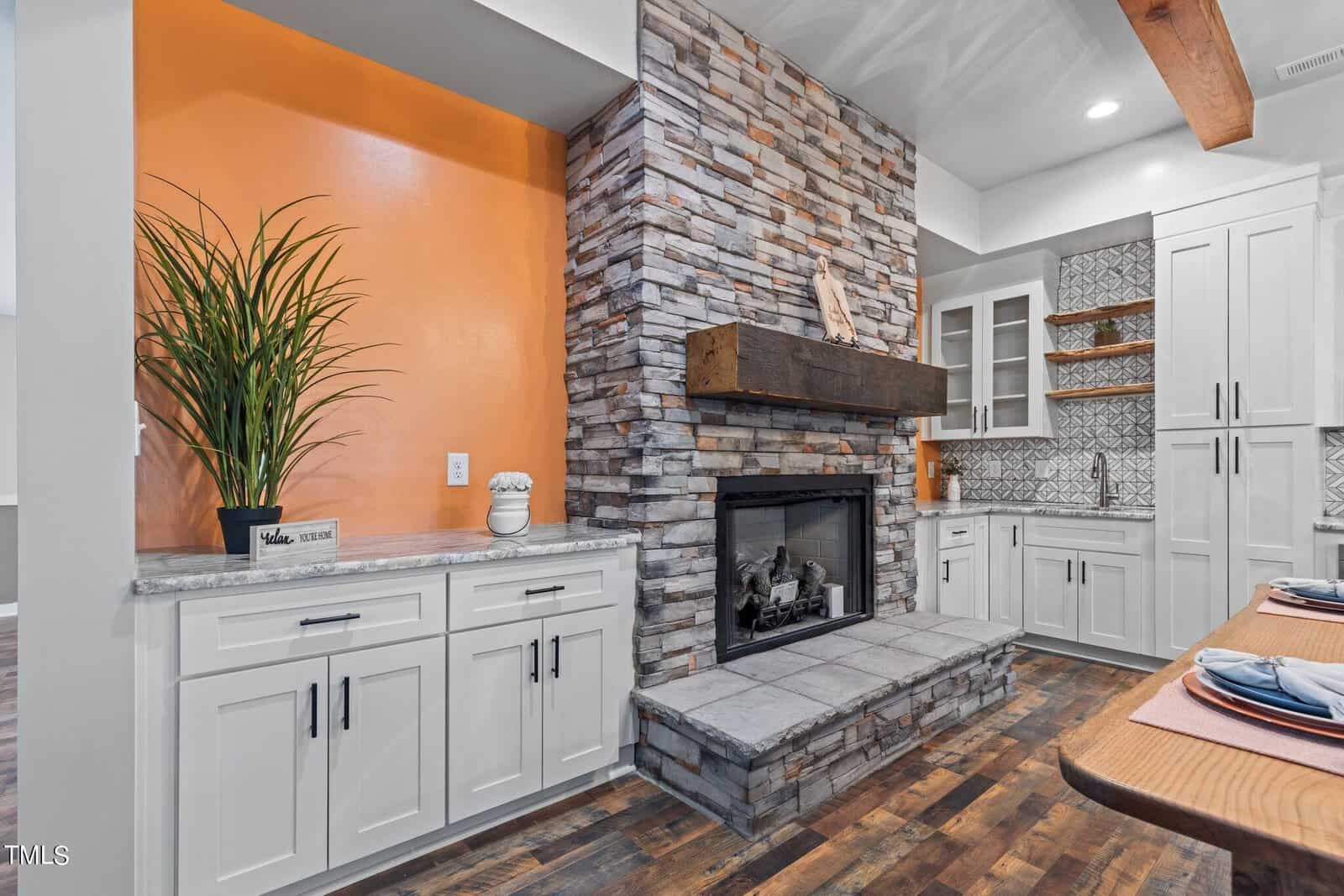 Modern kitchen with a stone fireplace, white cabinets, an orange accent wall, potted plant, patterned backsplash, and wooden floors, blending rustic and contemporary design elements.