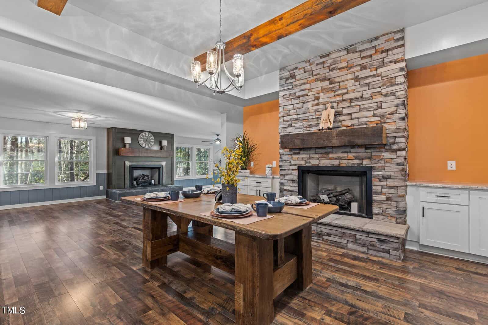 Rustic dining room with a wooden table set for six, stone fireplace with wooden mantel, orange accent wall, hardwood floors, ceiling beams, and large windows letting in natural light.