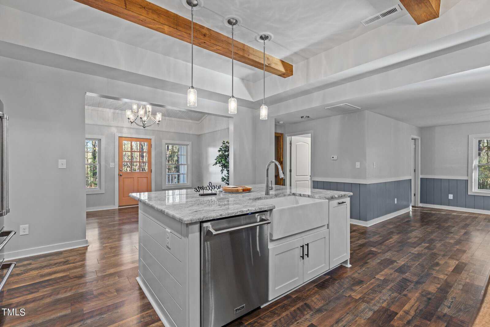 Modern kitchen with a marble island, farmhouse sink, pendant lights, and stainless-steel appliances. Open floor plan leads to a dining area with a chandelier and double front doors. Hardwood floors throughout.