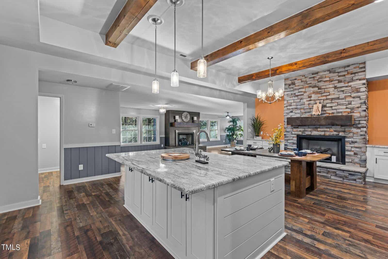 Modern kitchen with a large marble island, pendant lights, exposed wooden beams, hardwood floors, and a stone fireplace in the background next to a dining table; bright and spacious with white cabinets.