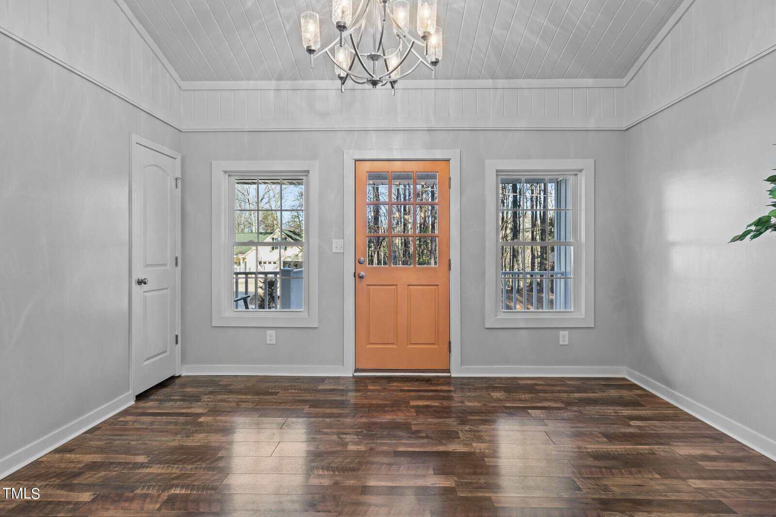 A bright, empty room with white walls, dark hardwood floors, two windows, a wooden front door with glass panes, a chandelier, and outdoor views through the windows.
