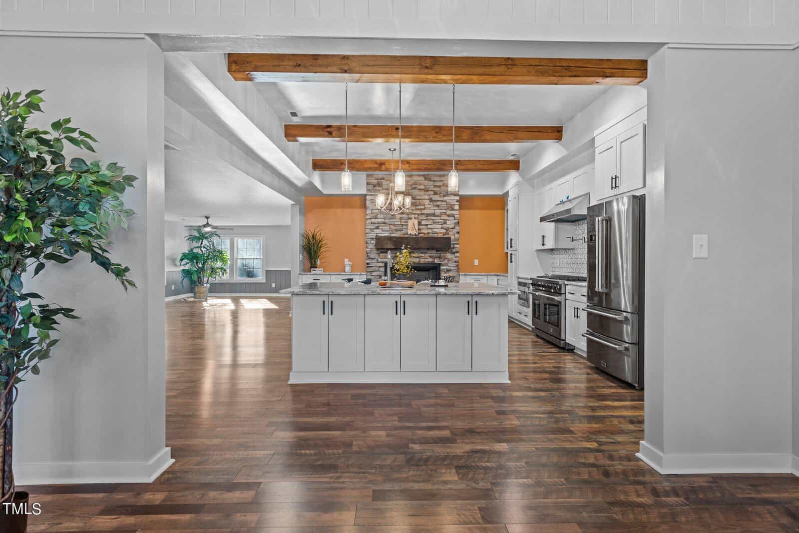 Open-concept kitchen with white cabinets, large island, stainless steel appliances, exposed wooden ceiling beams, and stone fireplace in the background. Hardwood floors and natural light fill the spacious area.