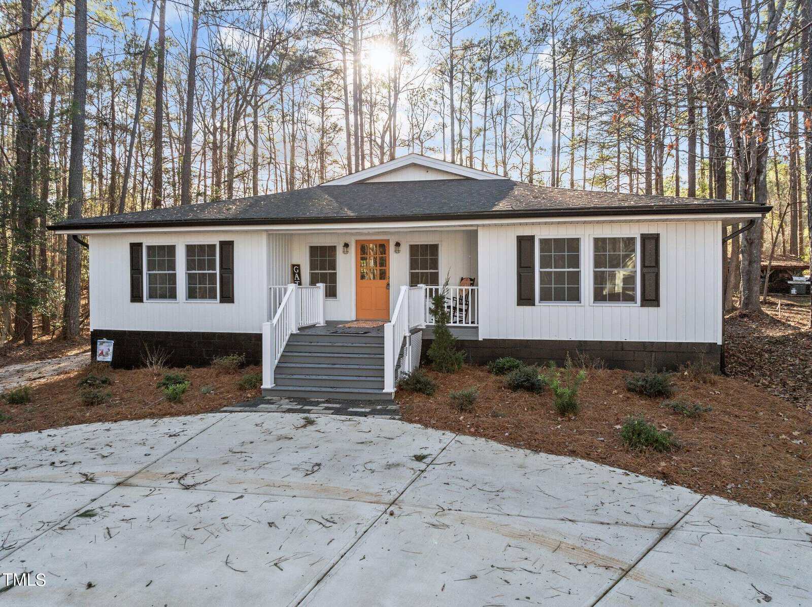 A white, single-story house with black shutters and a bright orange front door, surrounded by pine trees. There is a front porch with steps, simple landscaping, and a curved concrete driveway in front.