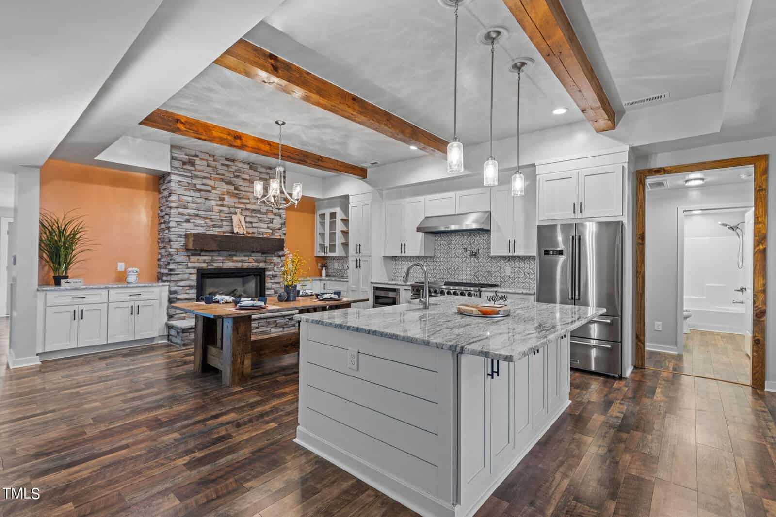 Modern kitchen with white cabinets, marble countertops, a large island, stainless steel appliances, exposed wood ceiling beams, wood flooring, and a stone fireplace in the background. Warm, inviting, and open layout.