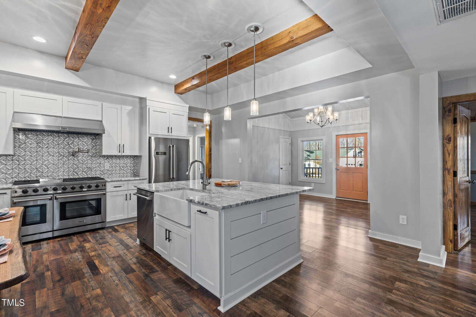 Modern kitchen with white cabinets, marble island, farmhouse sink, stainless steel appliances, patterned tile backsplash, pendant lights, wooden beams, and dark hardwood floors, opens to a hallway with a chandelier and orange front door.