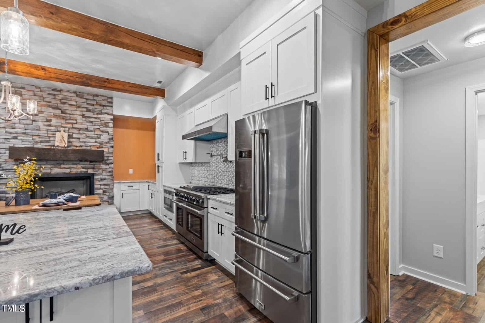 Modern kitchen with white cabinets, stainless steel appliances, and wood accents. An island with a granite countertop is in the foreground. The kitchen opens to a living area with a stone fireplace and wood beams on the ceiling.