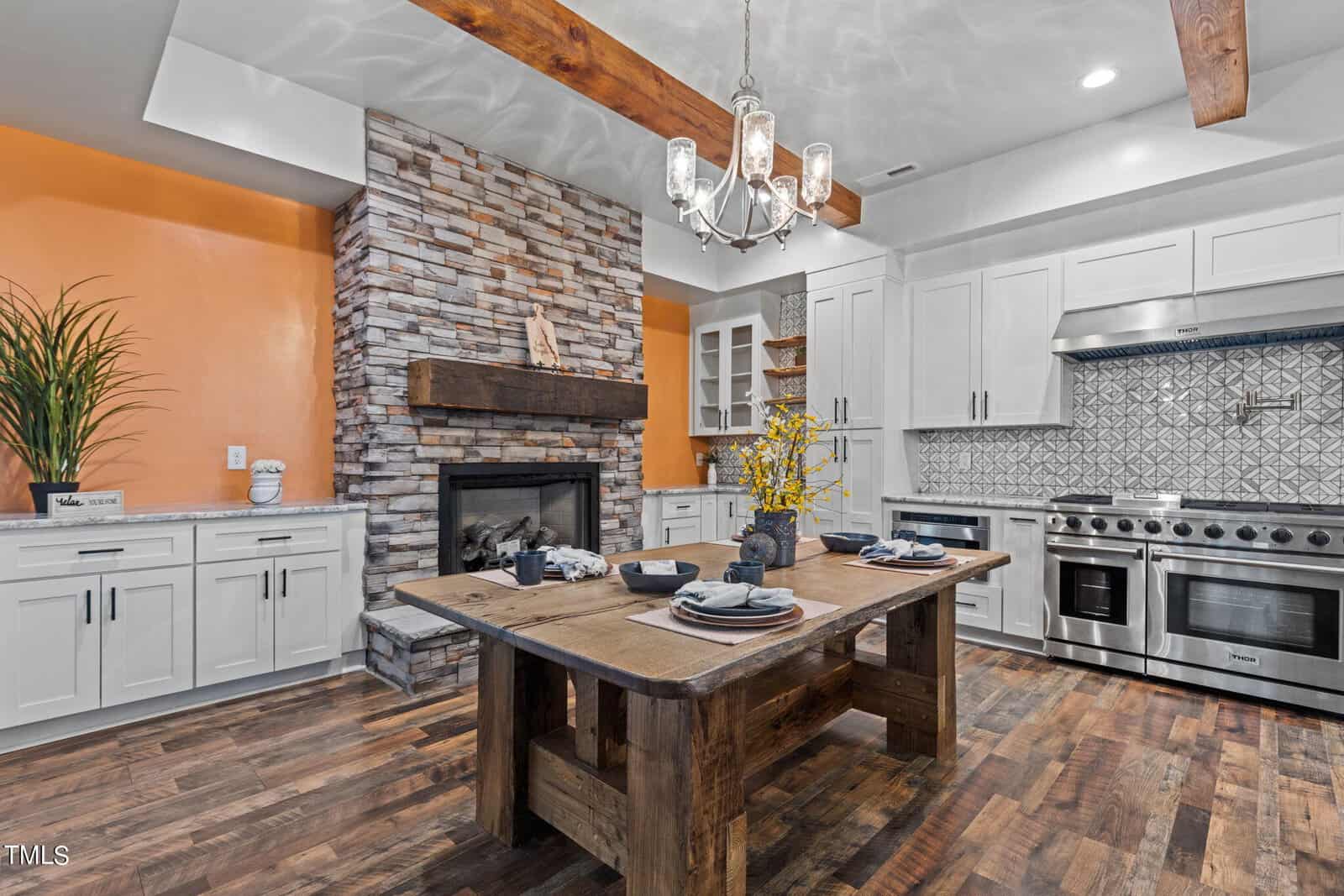A modern kitchen with white cabinets, stainless steel appliances, a patterned backsplash, a stone fireplace, wood beams, and a rustic wooden island set for dining. Orange accent wall and hardwood floors complete the space.