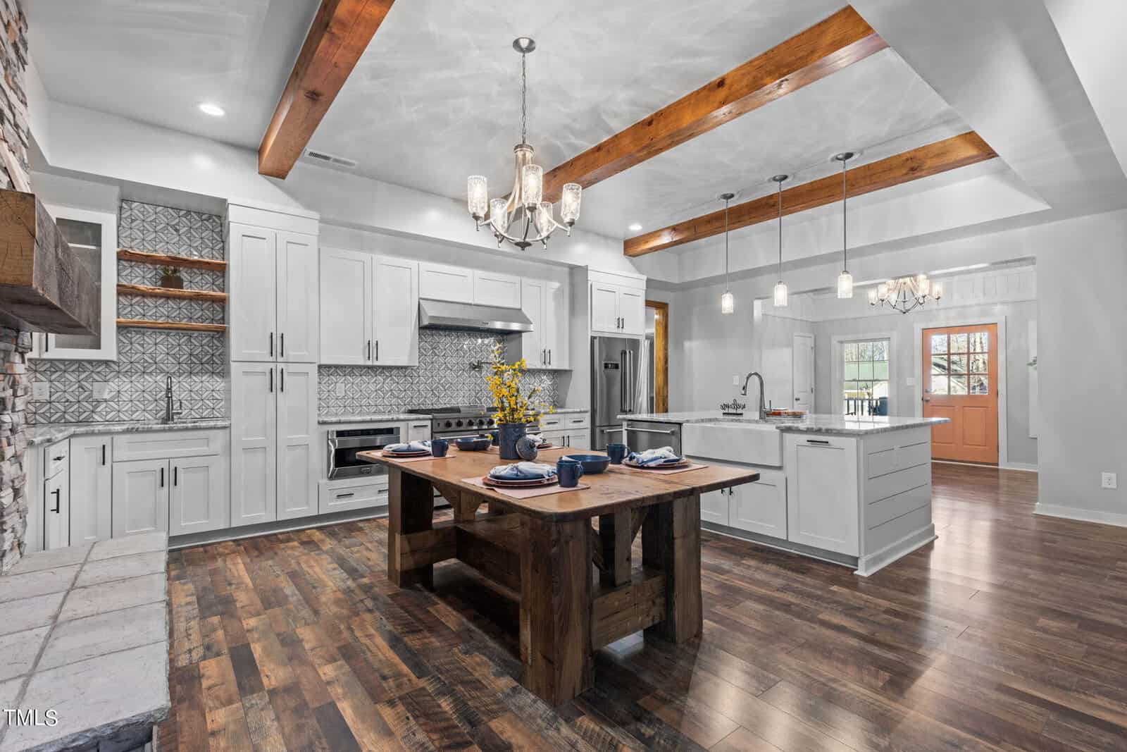 Modern farmhouse kitchen with white cabinets, wood beams, patterned backsplash, island, rustic dining table set for four, stainless steel appliances, and hardwood floors. Pendant and chandelier lighting add warmth to the space.