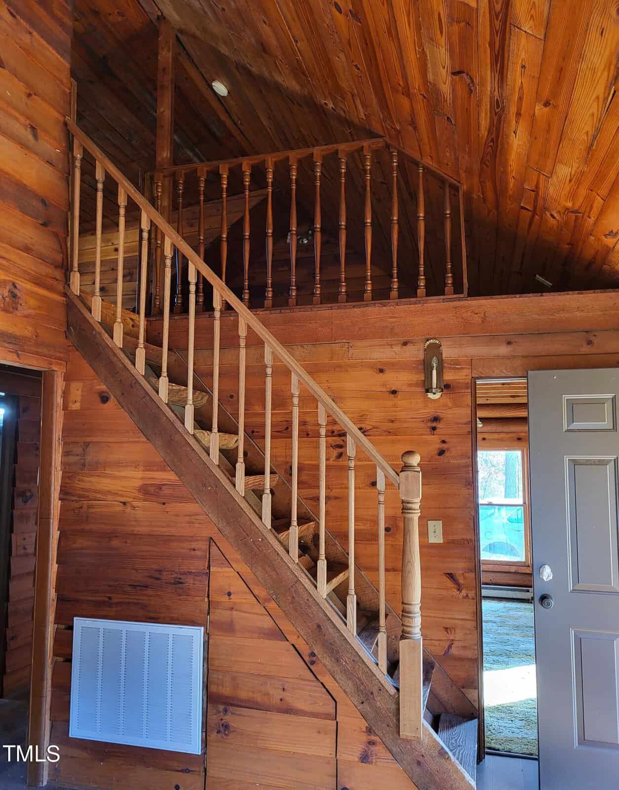 Wooden interior of a cabin featuring a staircase with a simple railing. The walls and ceiling are made of wood, and the open door leads to a room with carpet. A wall-mounted light fixture is visible beside the stairs.