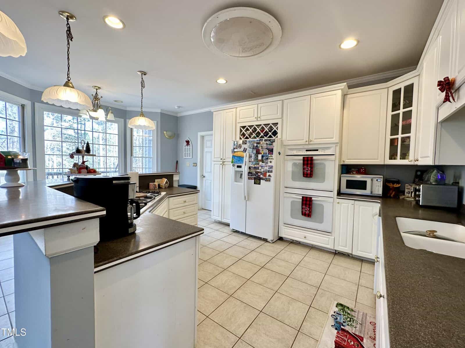 A spacious kitchen with white cabinets and countertops, featuring double ovens, a fridge with magnets, a microwave, and pendant lights. Large windows provide natural light. A small island and bar stools are visible in the foreground.