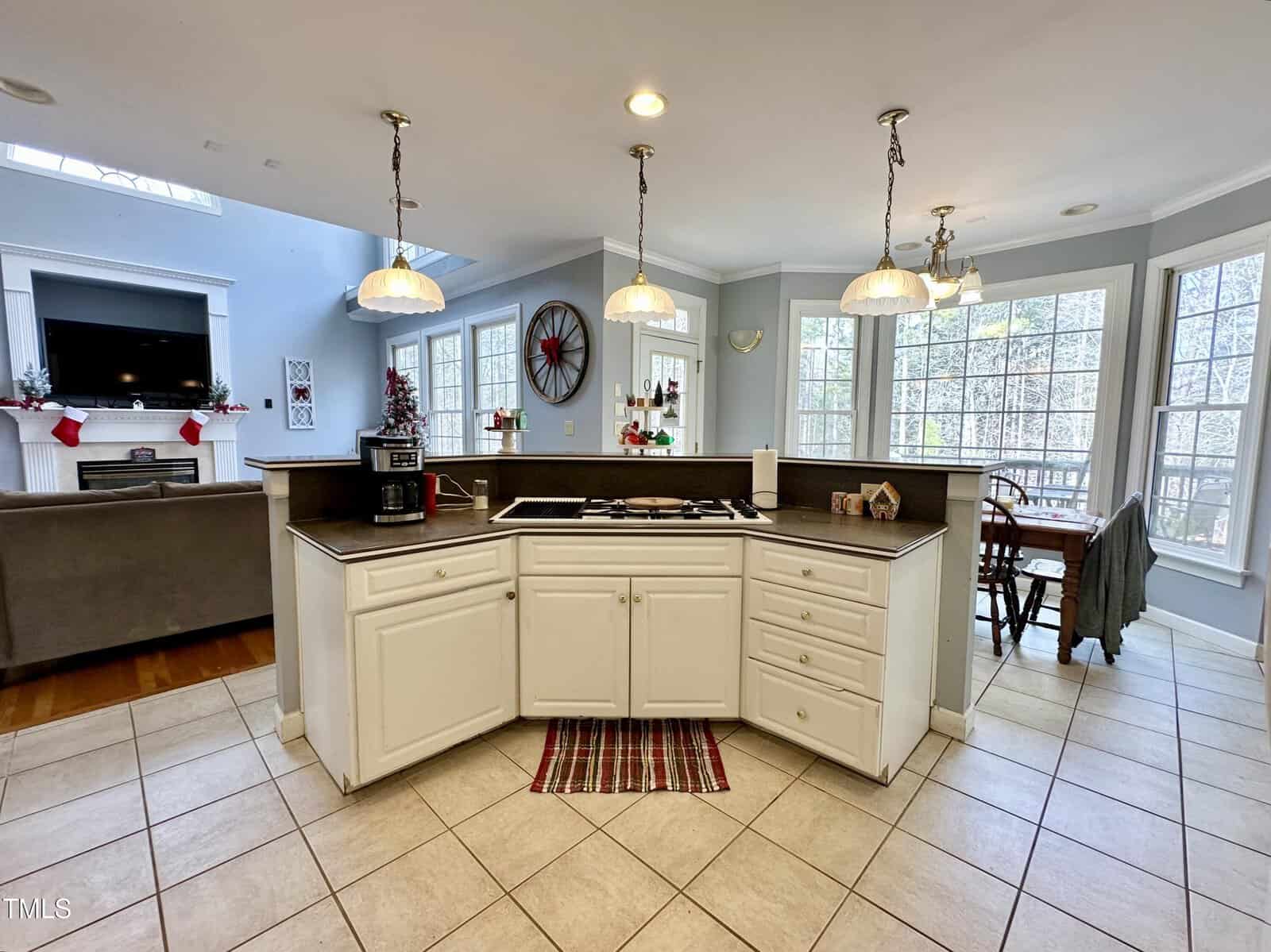 A spacious kitchen with white cabinets and a central island featuring a cooktop and soap dispenser. Large windows let in natural light. A dining table is visible in the background, and a living area with a fireplace is on the left.
