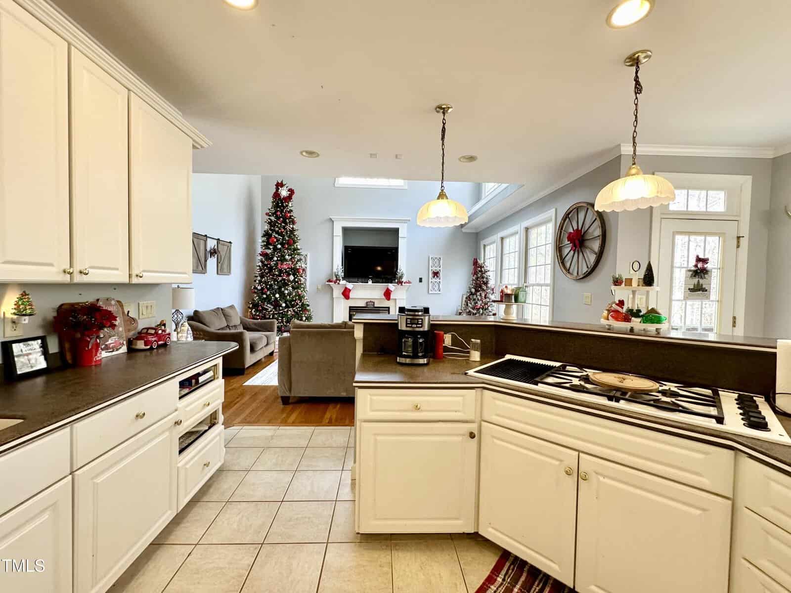 A spacious kitchen and living room area decorated for Christmas. The kitchen has white cabinets and a dark countertop, with a stove and hanging lights. In the background, there's a Christmas tree and a cozy seating arrangement near a fireplace.