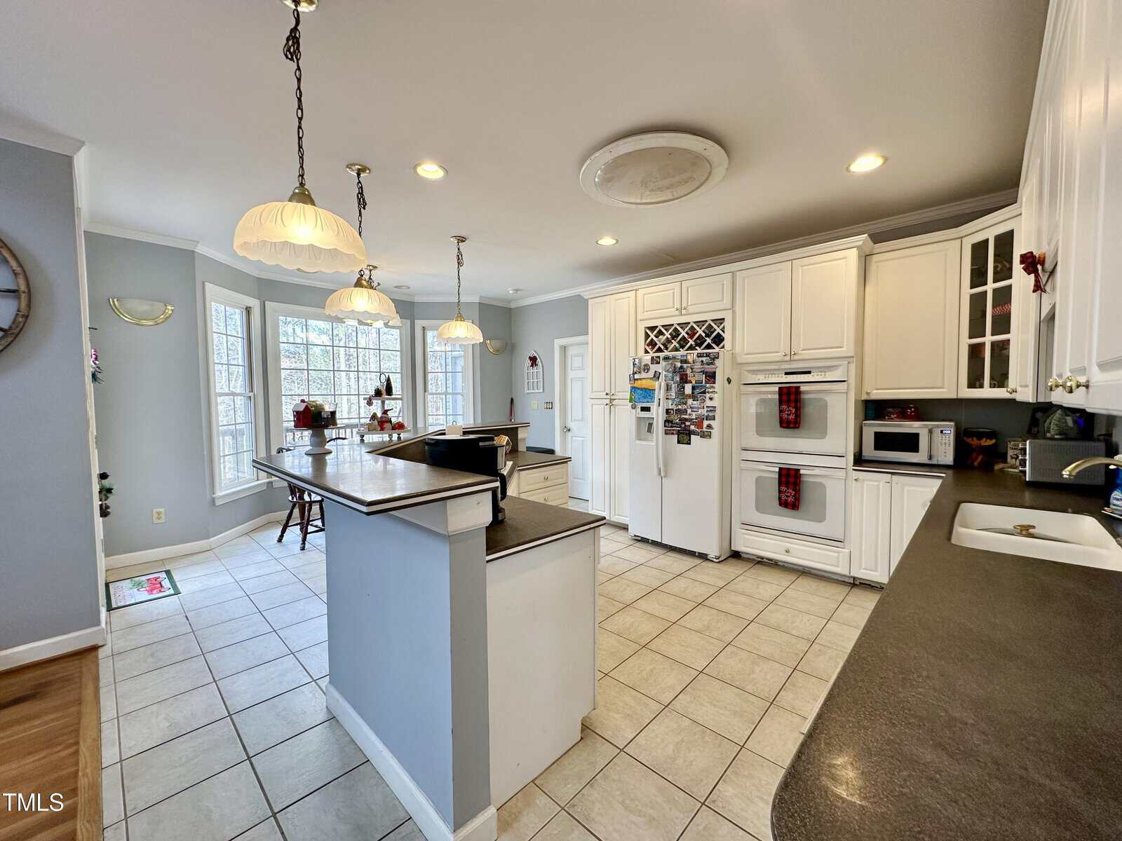 Spacious kitchen with white cabinetry, a central island with granite countertops, and pendant lighting. Large windows allow natural light, and modern appliances include a stove, double oven, and refrigerator. Tile flooring completes the look.