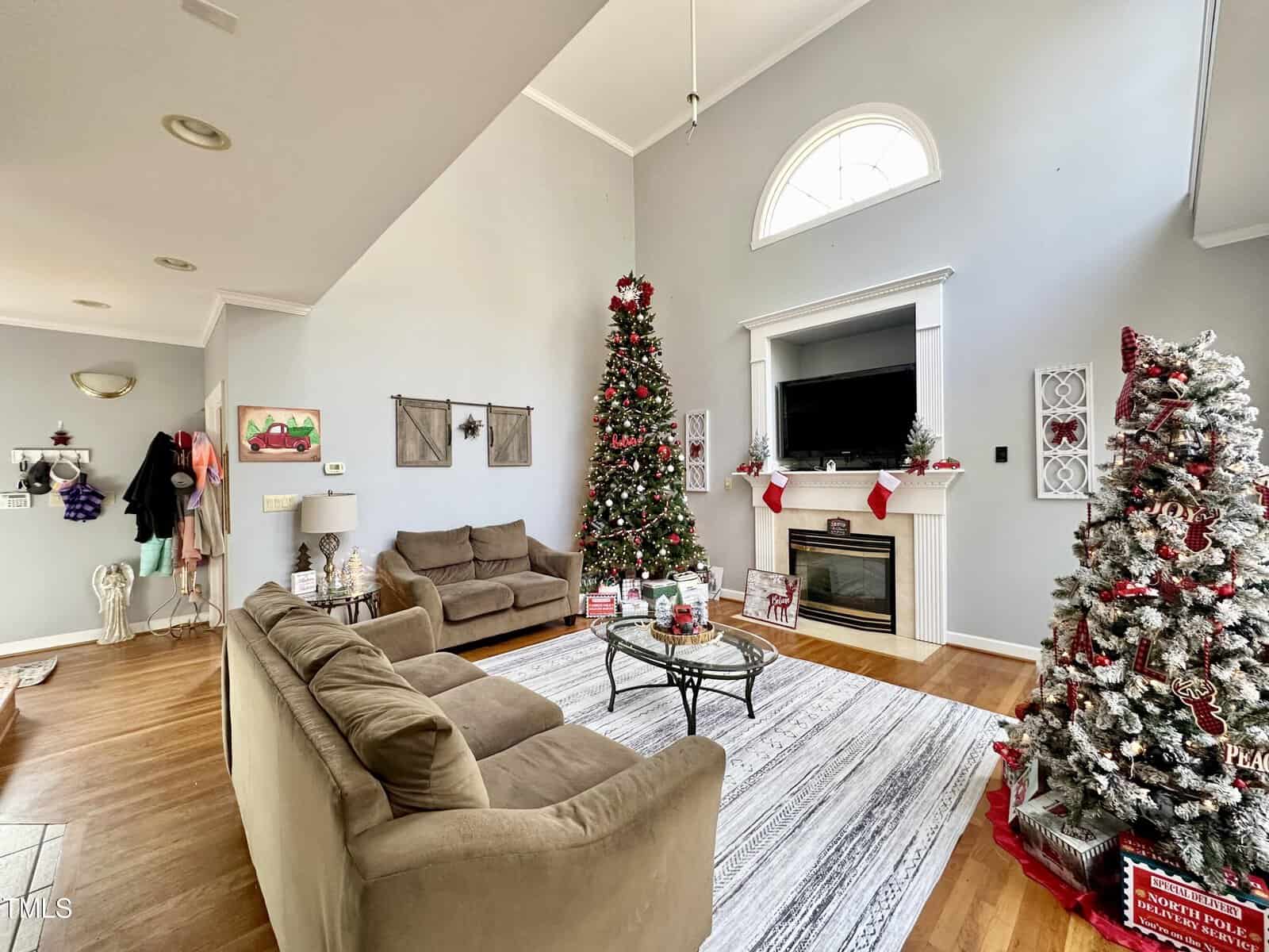 A spacious living room with high ceilings features two decorated Christmas trees, a fireplace with a TV above it, and cozy brown sofas on a striped rug. Holiday decorations, including stockings and wreaths, add festive charm to the space.