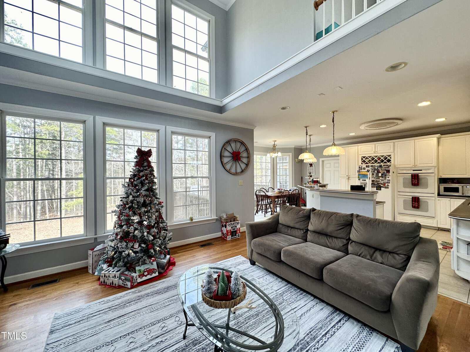 A spacious, bright living room with large windows and a high ceiling. There's a decorated Christmas tree, a gray sofa, and a glass coffee table on a patterned rug. The open kitchen in the background features white cabinets and bar seating.