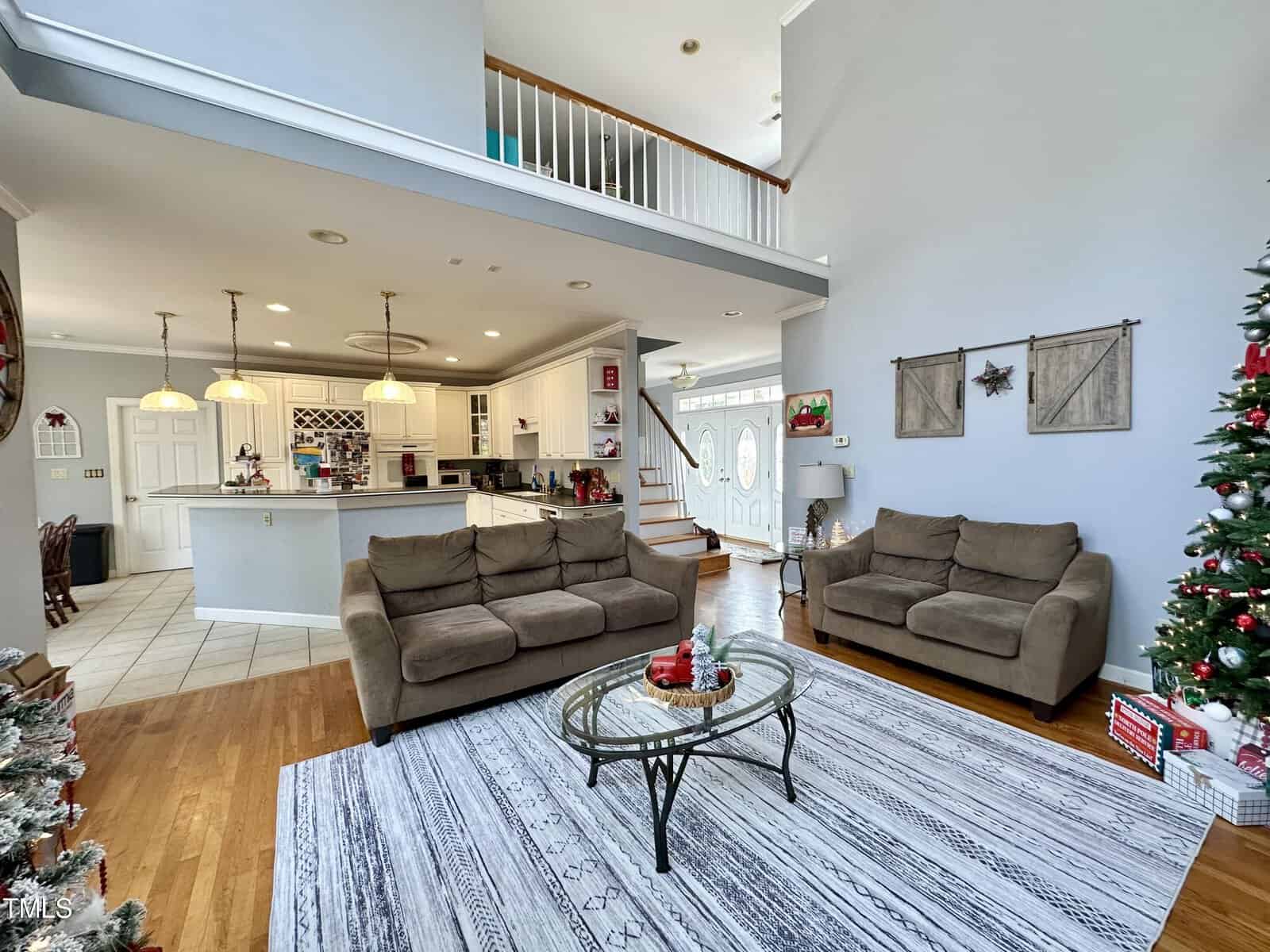 A spacious living room with high ceilings, featuring two brown couches, a glass coffee table, and a Christmas tree. The open layout extends to a kitchen with white cabinets, pendant lights, and bar stools. A staircase leads to an upper level.