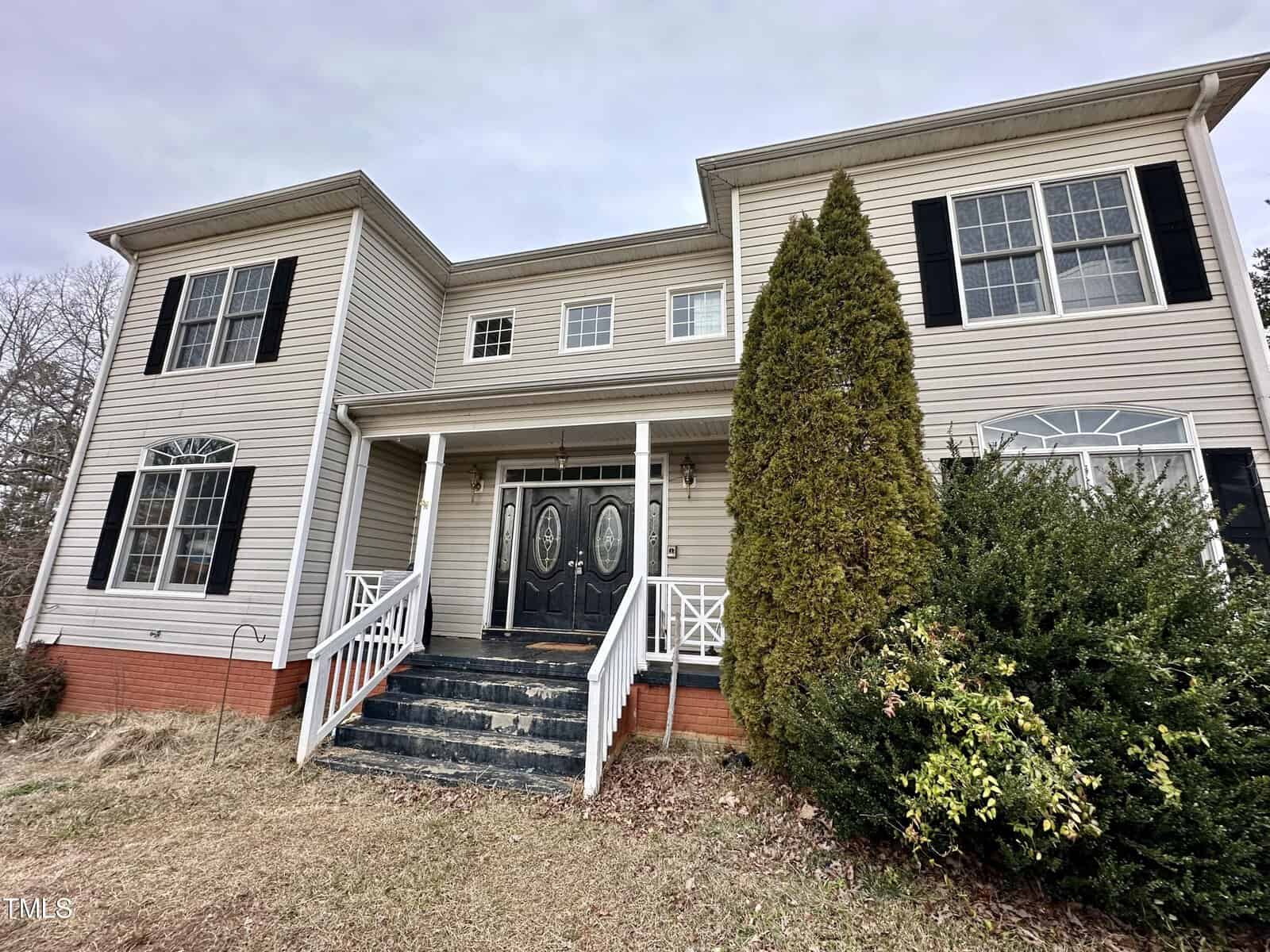 A two-story house on Oxford's 7680 Us-15 boasts beige siding and black shutters, complete with a welcoming front porch featuring double doors and stairs. The grassy yard is adorned with a large bush and tree near the entrance, set against an overcast sky.