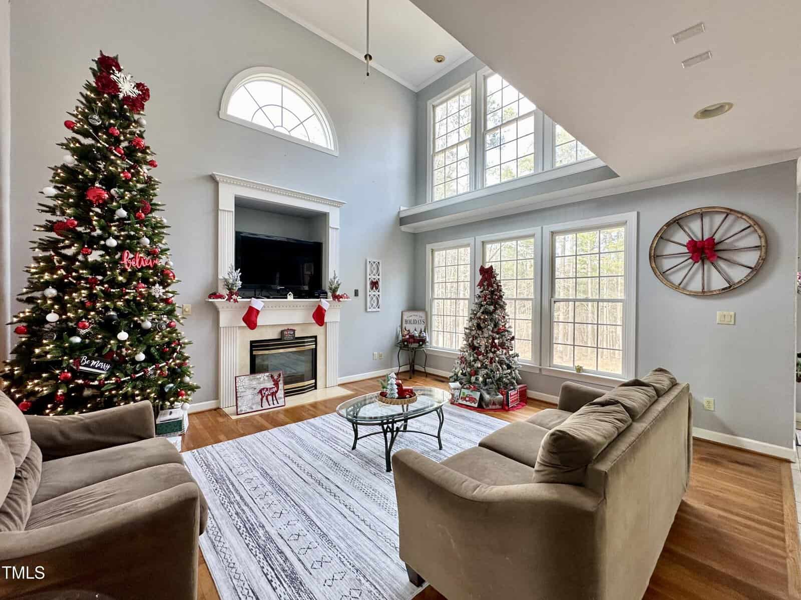 A cozy living room adorned for Christmas, featuring two decorated Christmas trees, a fireplace with stockings, a TV above the mantel, and large windows letting in natural light. Two plush sofas and a patterned rug complete the scene.