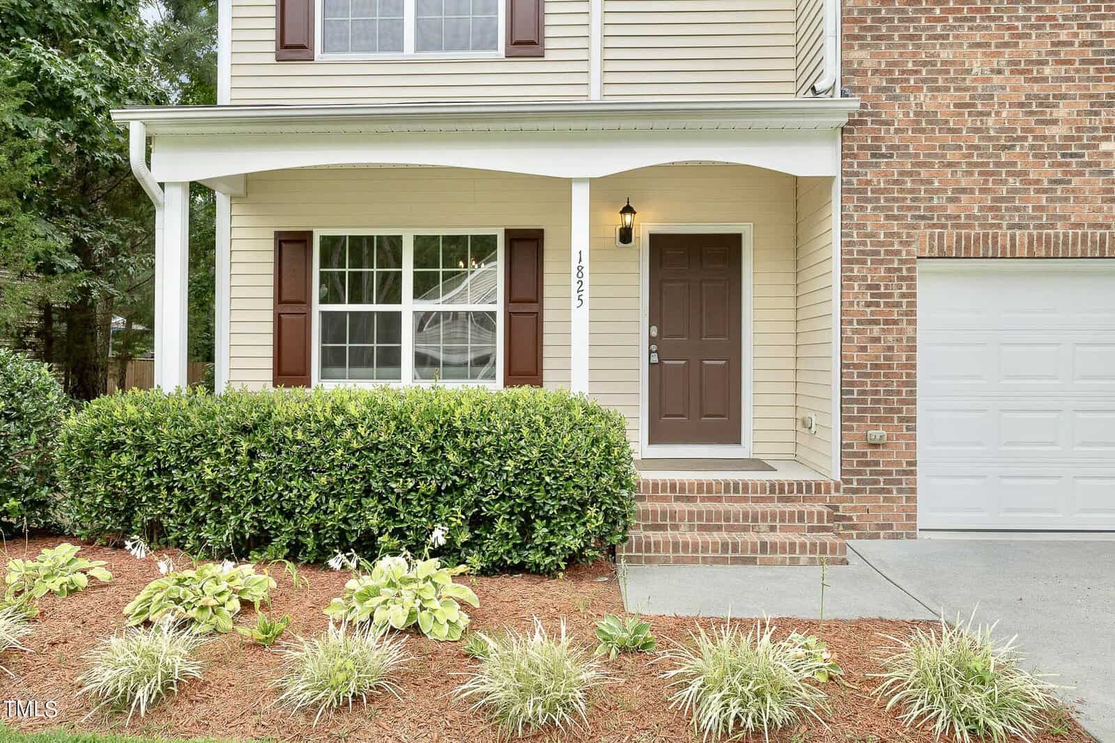 Front view of a two-story house with beige siding and brick accents. There are white pillars, a dark brown front door, a large window with shutters, and a garage. The front garden has shrubs and small plants.