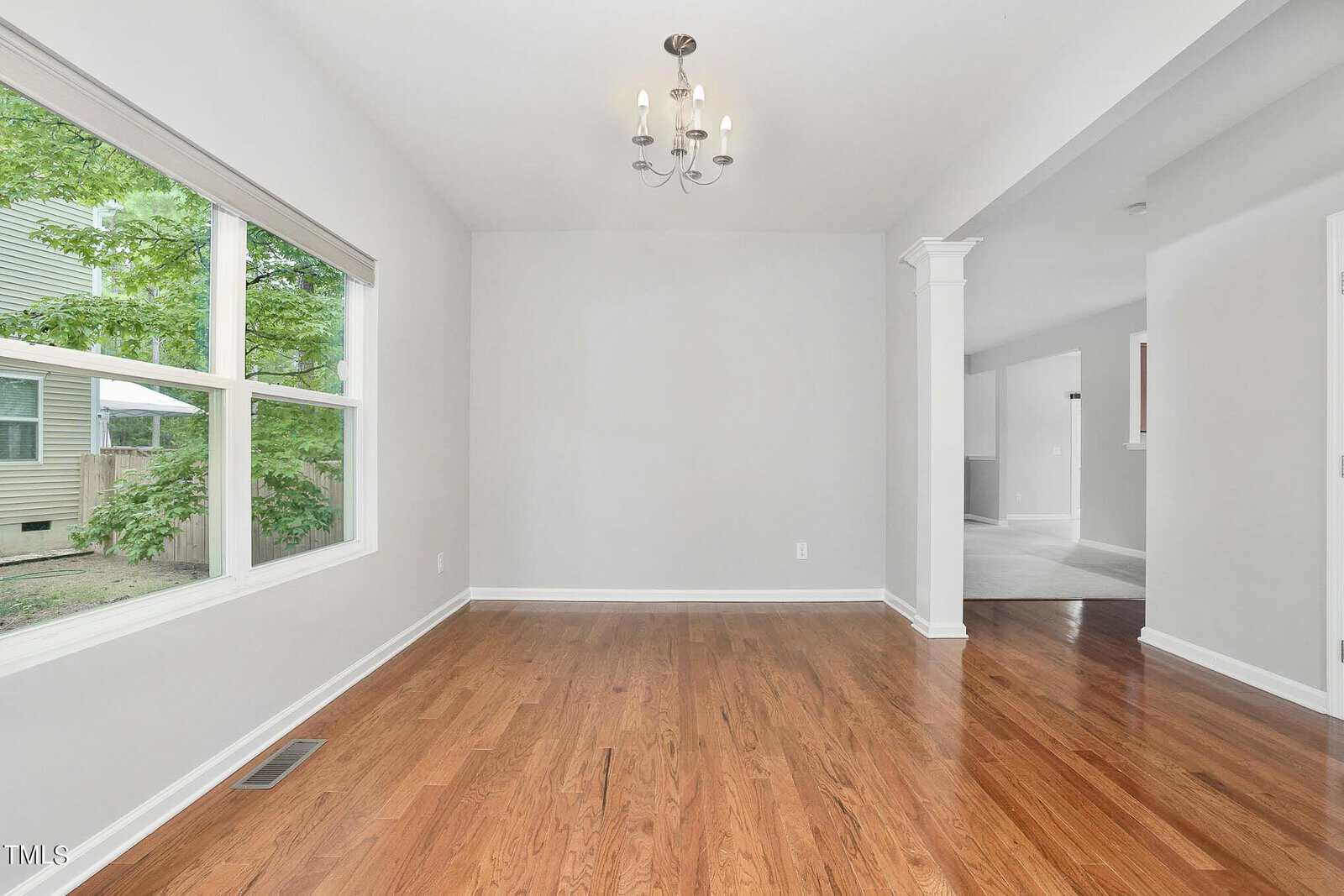 An empty room with hardwood floors, a large window on the left revealing greenery outside, a small chandelier hanging from the ceiling, white walls, and an open doorway leading to another room with carpet.