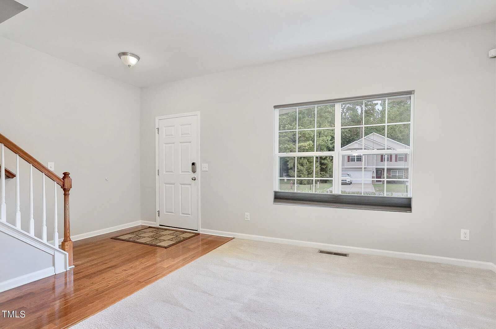Bright and spacious living room with beige walls and carpet. A wooden staircase is on the left, and a window on the right offers a view of a house and greenery outside. A white door with a small mat is near the stairs.