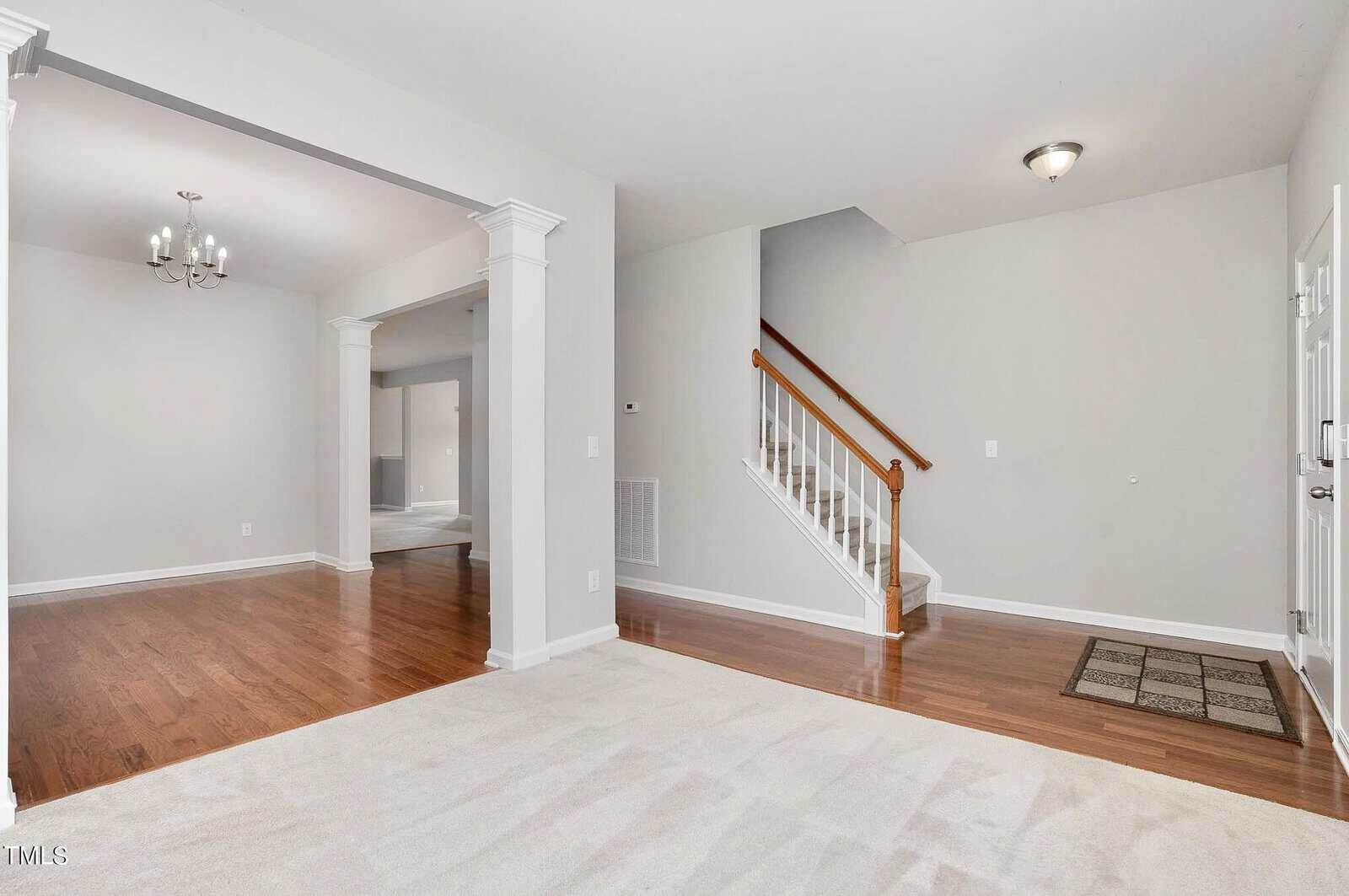 Image showing a bright, empty interior space with light gray walls and hardwood floors. In the foreground is a staircase with wooden steps and a white banister. A chandelier hangs in the adjoining dining area, visible through an open archway.