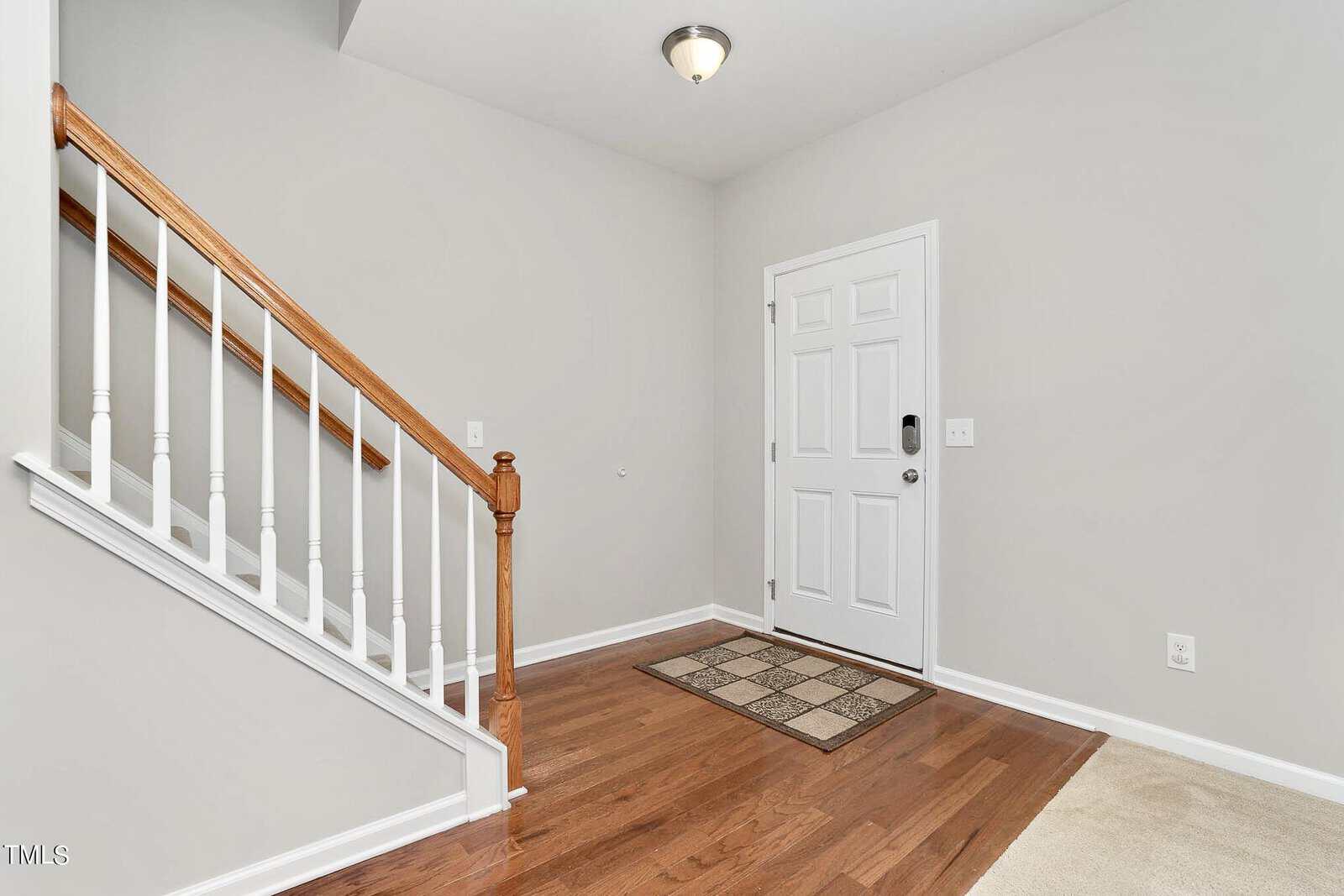 A minimalist entryway featuring a wooden staircase with white balusters and a white door. The hardwood floor contrasts with a beige carpeted area. A small geometric rug lies by the door, and a single ceiling light hangs above.