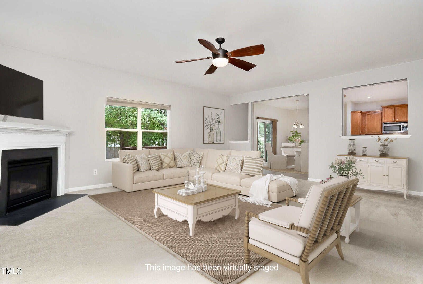 A virtually staged living room featuring a beige sectional sofa, a wooden coffee table, and a fireplace with a flat-screen TV above it. The room has large windows, a ceiling fan, and a view into a dining area and kitchen.