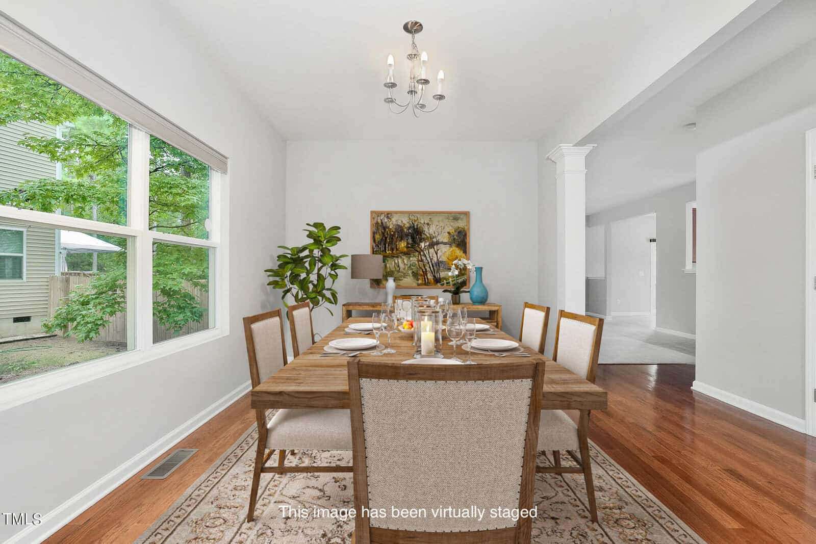 A virtually staged dining room featuring a wooden table set for six with white cushioned chairs. A chandelier hangs above, and a plant and painting decorate the back wall. Large windows let in natural light, revealing green foliage outside.