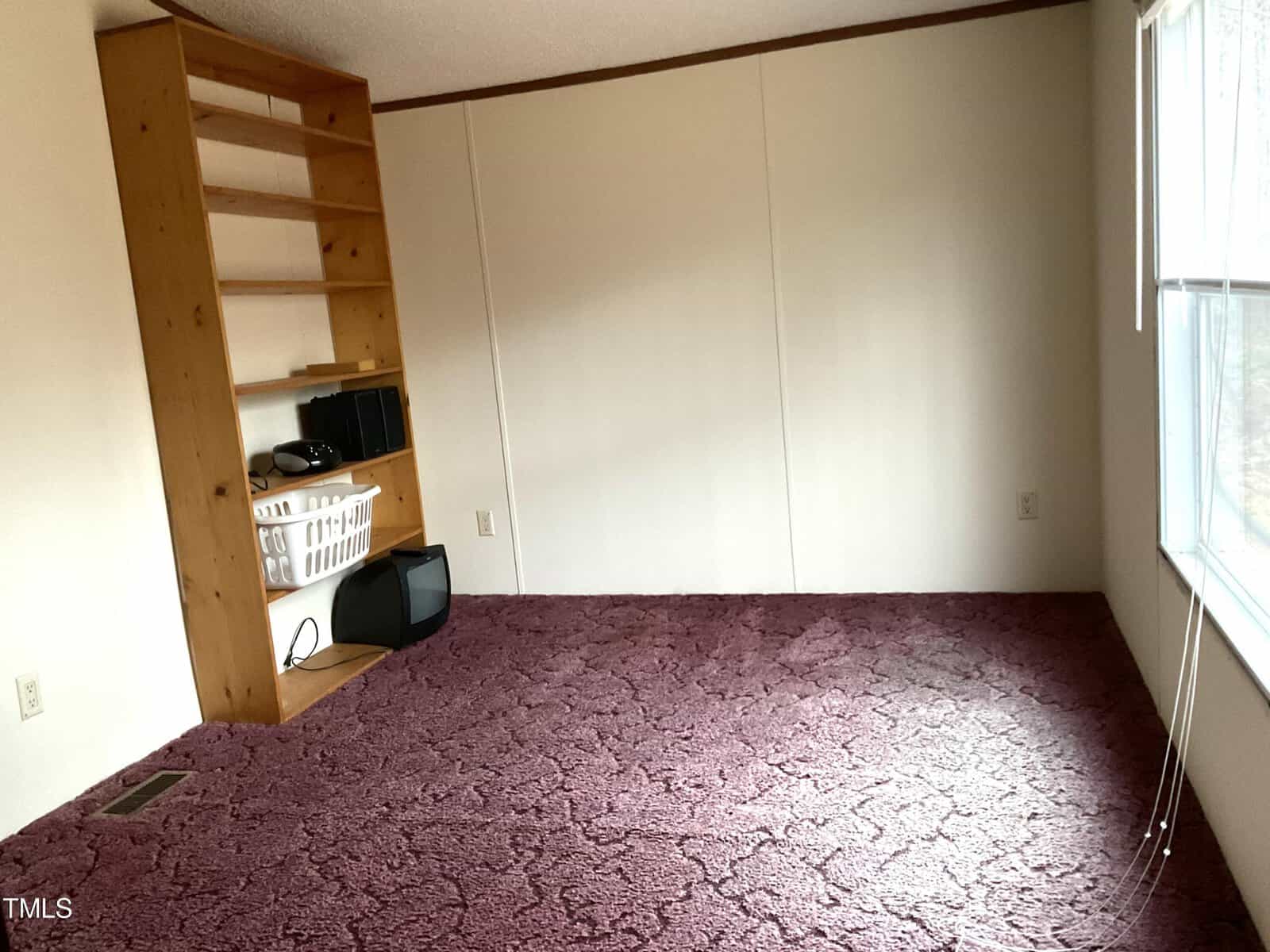 A sparsely furnished room with a tall wooden bookshelf in the corner holding electronics and a laundry basket. The floor is covered with dark red carpet, and a large window on the right lets in natural light. The walls are plain and off-white.