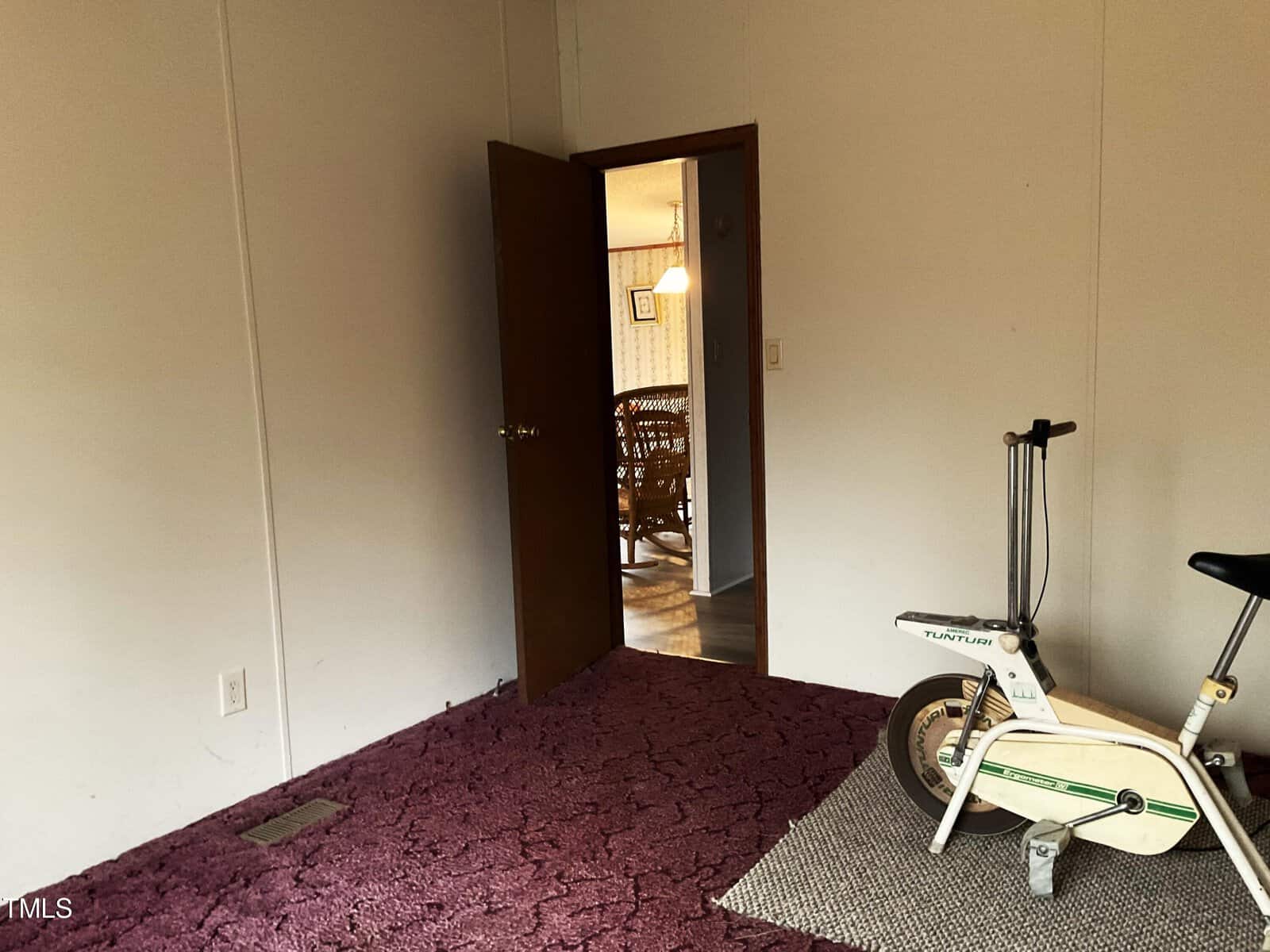 Room with maroon carpet and an open door leading to a dining area. A vintage exercise bike is on a small gray rug in the corner. Soft light from the adjacent room highlights a wooden dining table and chairs.