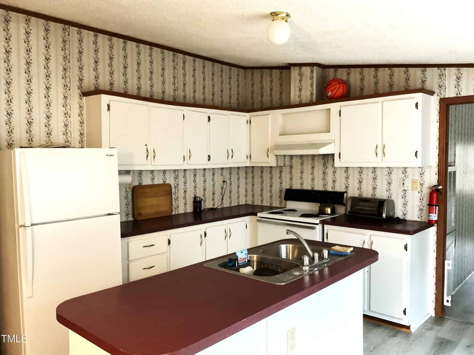 A cozy kitchen with white cabinets, a red countertop, and floral wallpaper. It features a refrigerator, electric stove, sink, and various kitchen items. A basketball and fire extinguisher are visible on top of the cabinets and in the corner, respectively.