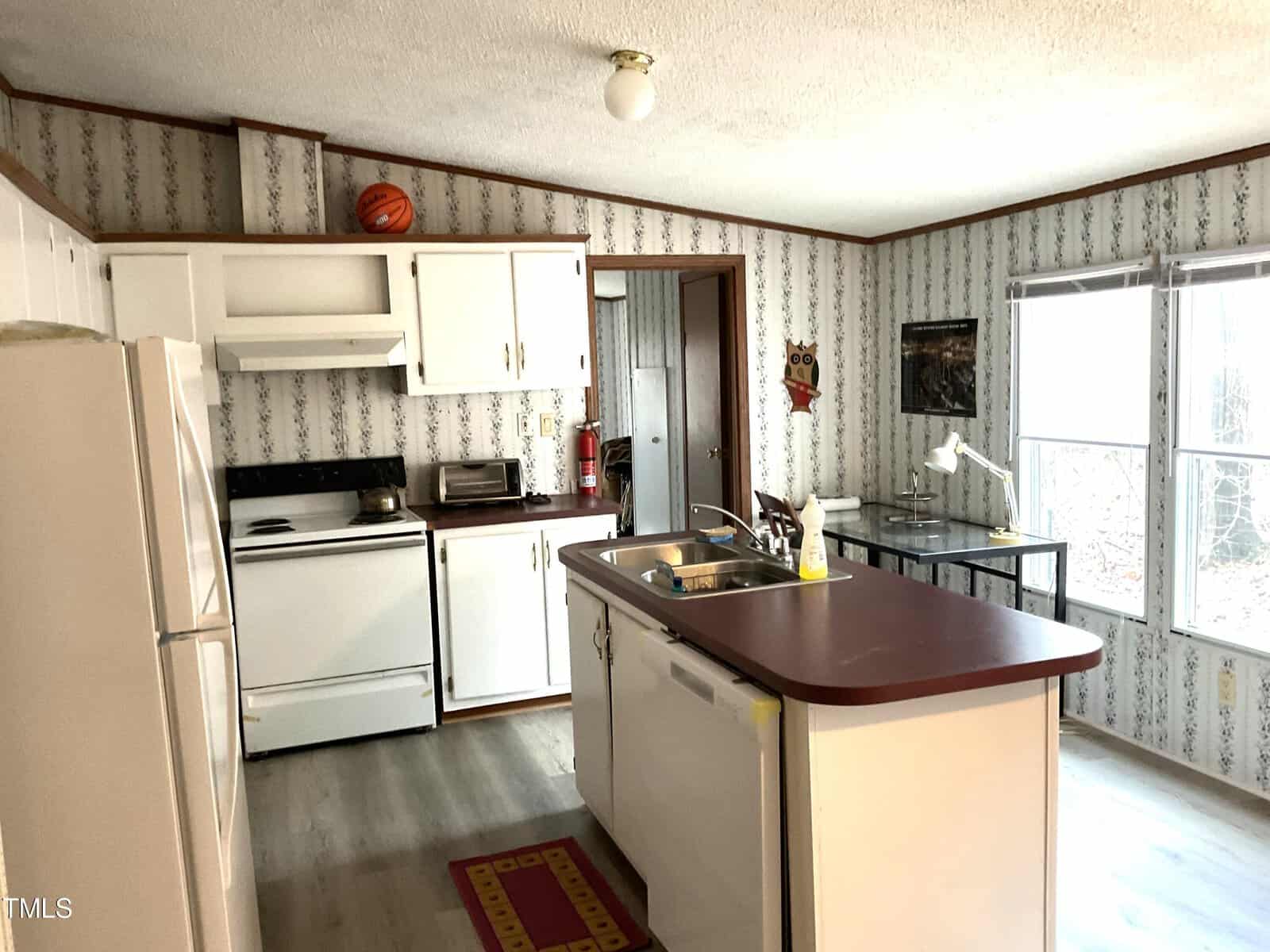 A cozy kitchen featuring white cabinets, a maroon countertop island with a sink, a white fridge, and a stovetop oven. A basketball sits atop the cabinets. Large windows provide natural light, and the walls are covered in floral wallpaper.