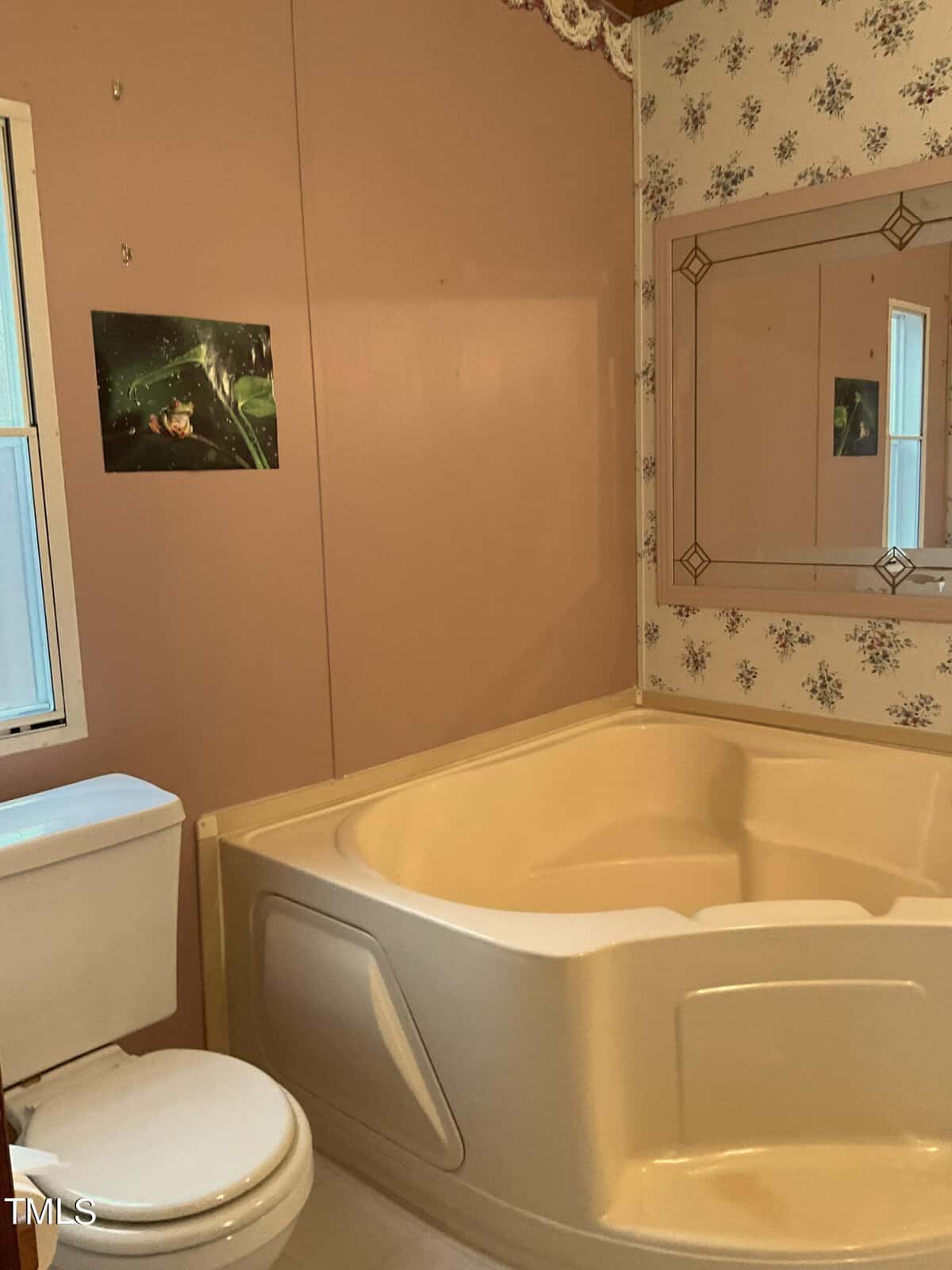 A bathroom featuring a corner bathtub beside a toilet. The walls are a mix of floral wallpaper and solid paint. A rectangular mirror hangs above the bathtub, reflecting a window. A small framed picture is on the wall above the toilet.