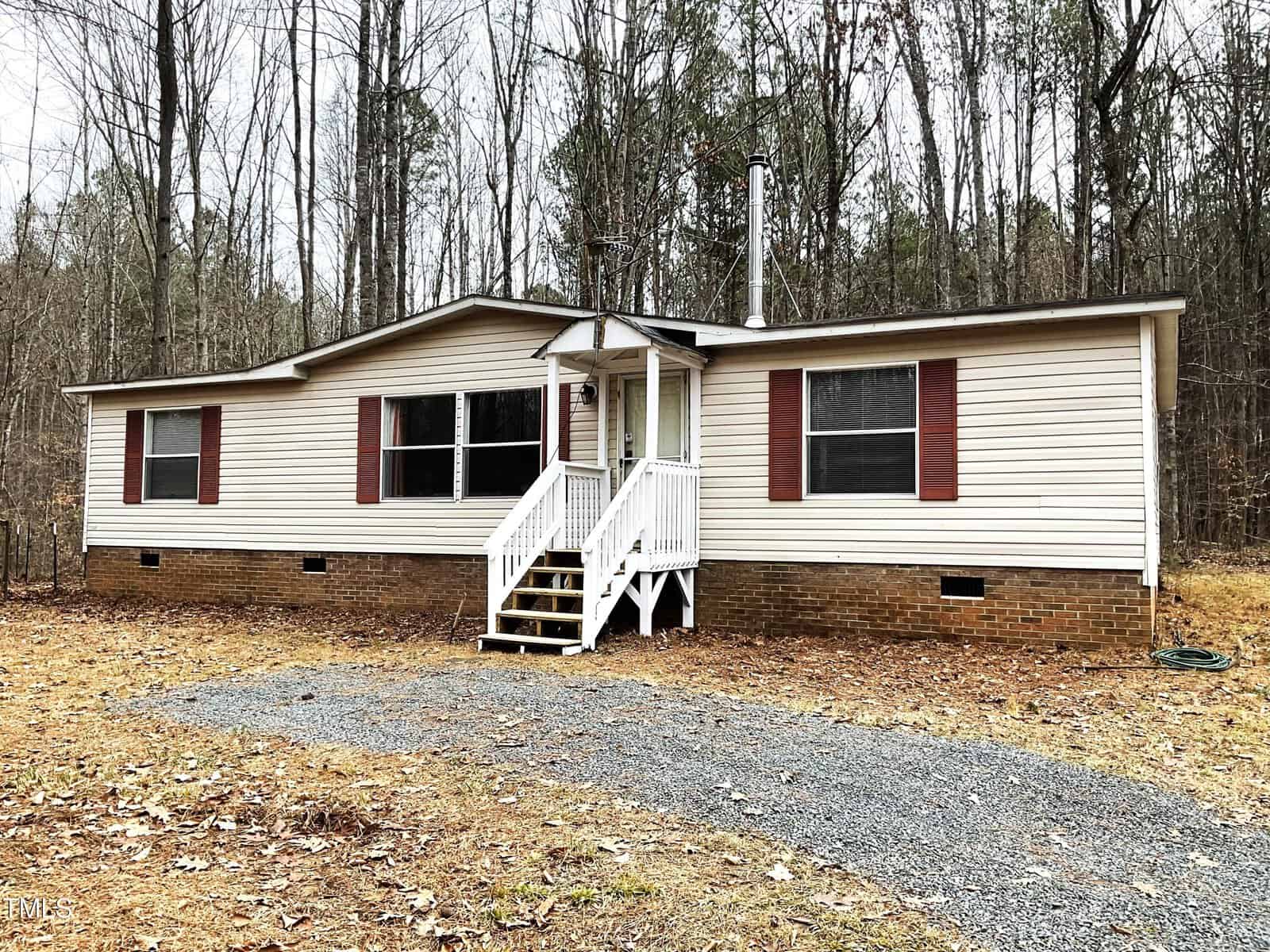 A beige manufactured home with a brick foundation sits in a wooded area. It has maroon shutters, a front porch with white railings, and steps leading to a gravel path. The surrounding trees have sparse leaves, indicating a fall or winter season.