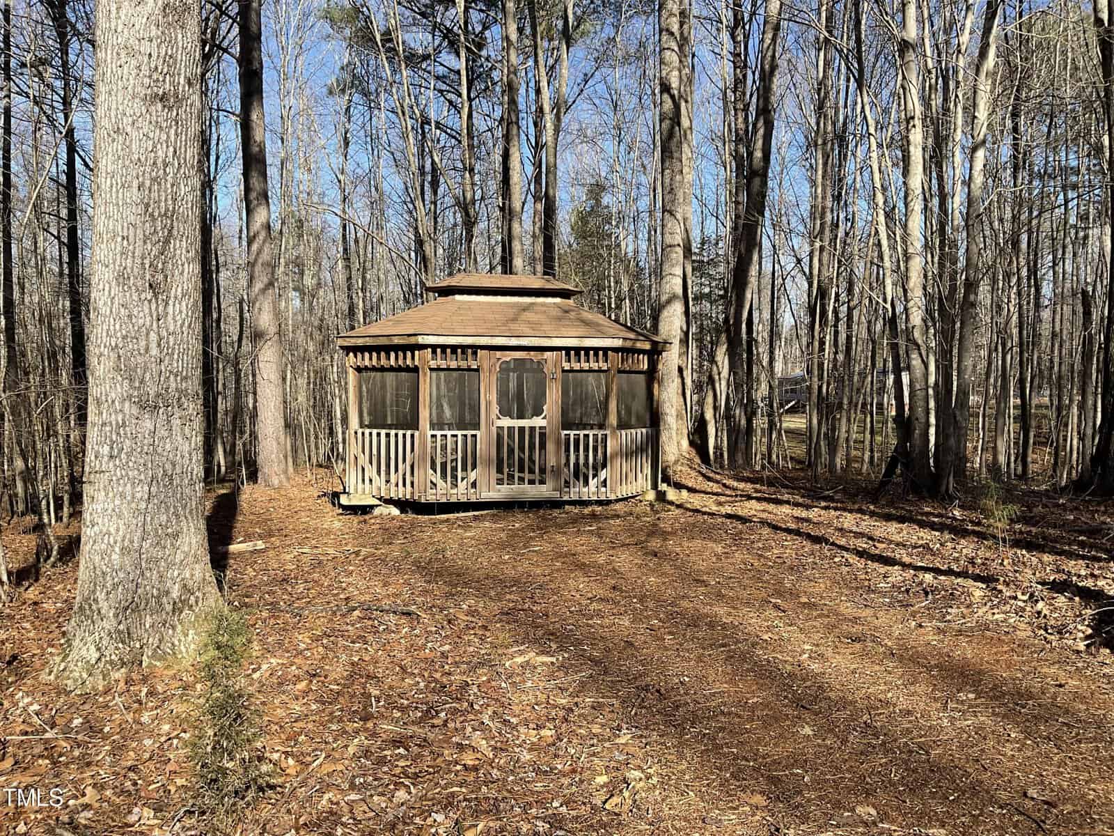 A small wooden gazebo with screen windows and a porch stands in a wooded area. The ground is covered with dry leaves, and tall trees surround the structure. The sky is clear, indicating a sunny day.