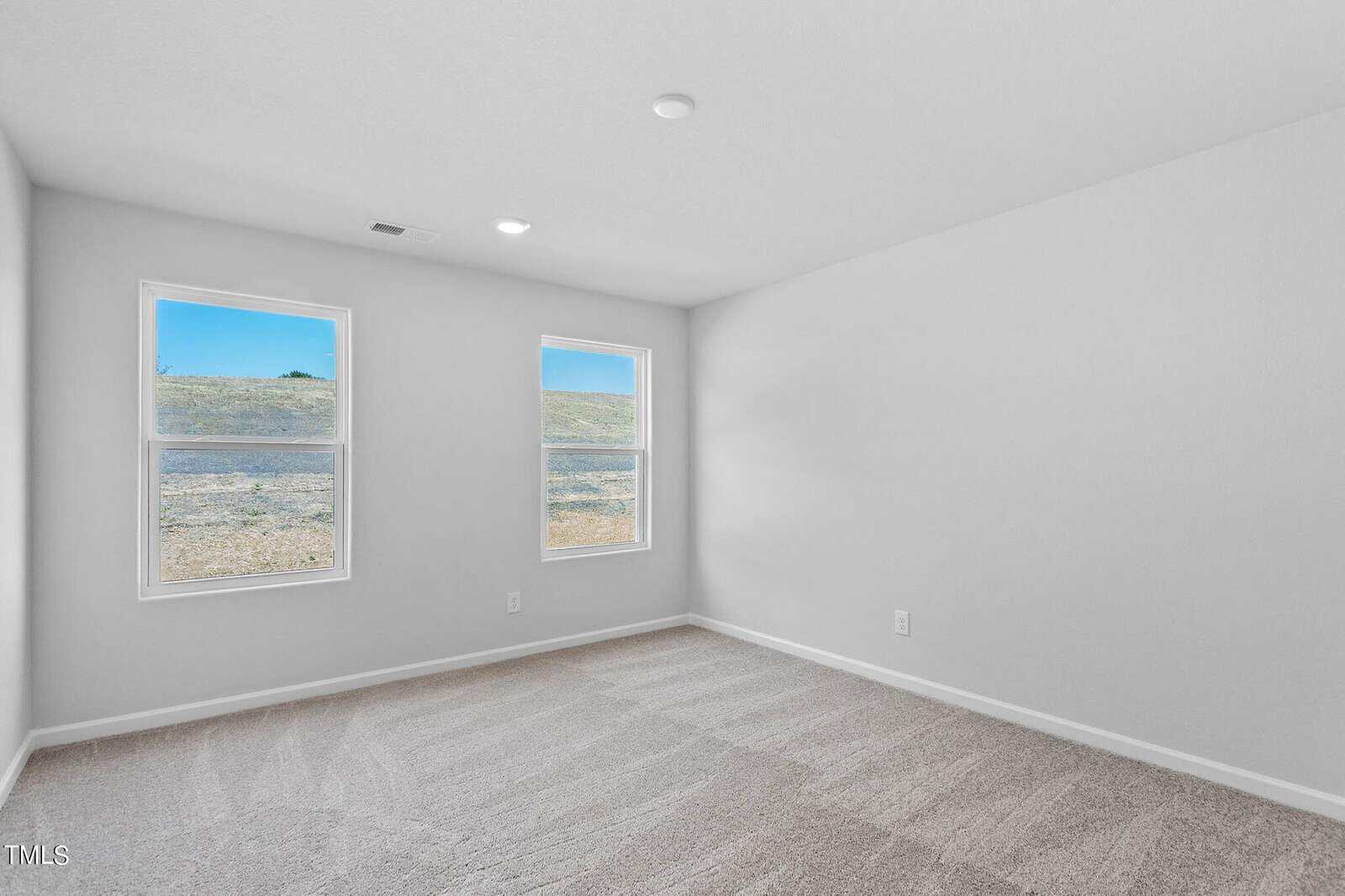 An empty room with light gray walls and two windows showing a grassy landscape. The floor is carpeted in a beige texture, and the room has a simple ceiling light fixture.