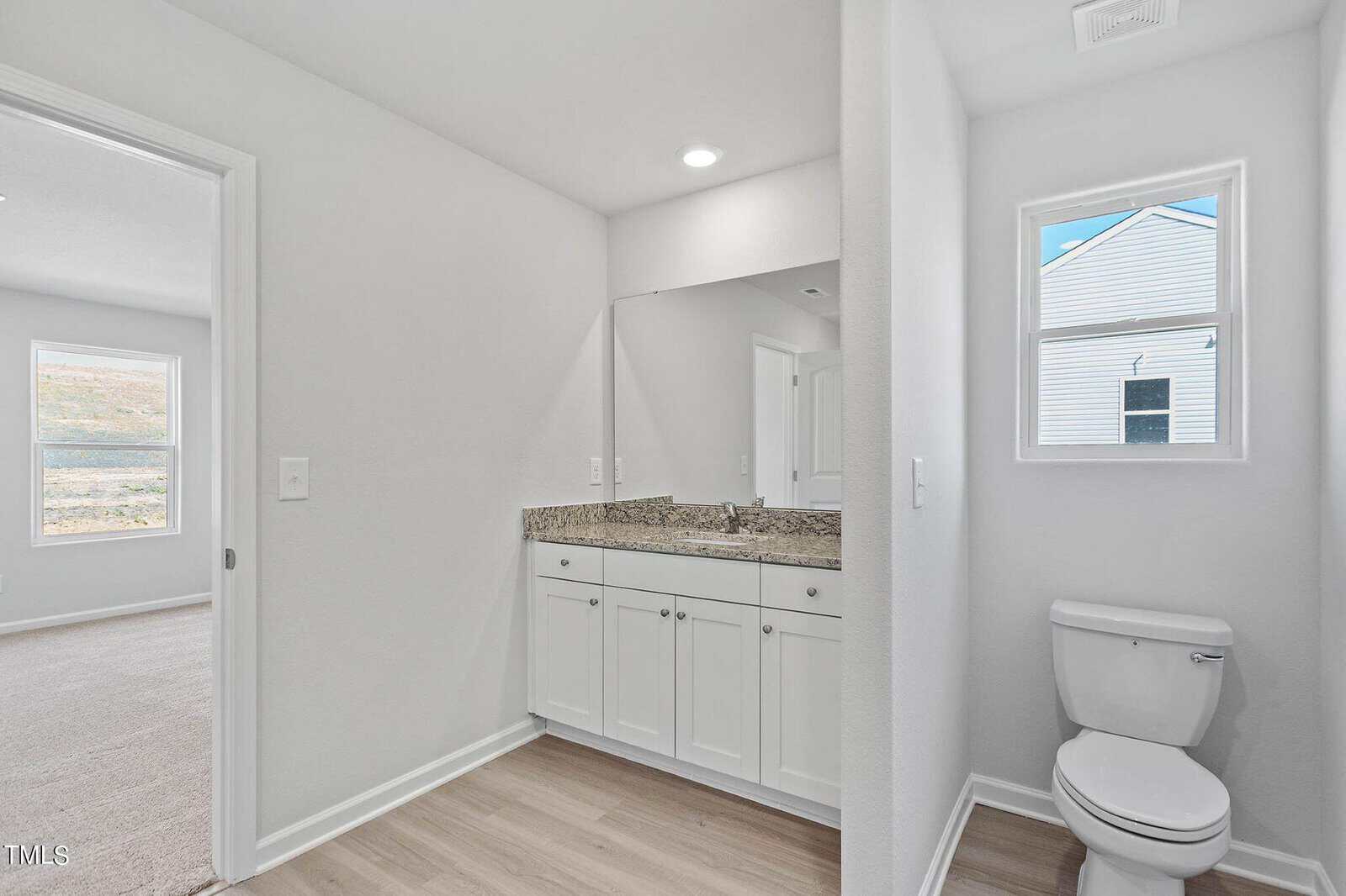 A bathroom with white walls and light wood flooring. It features a granite countertop with a large mirror above a white vanity. A toilet is positioned near a small window, with a view of a neighboring house outside. A doorway leads to a carpeted room.