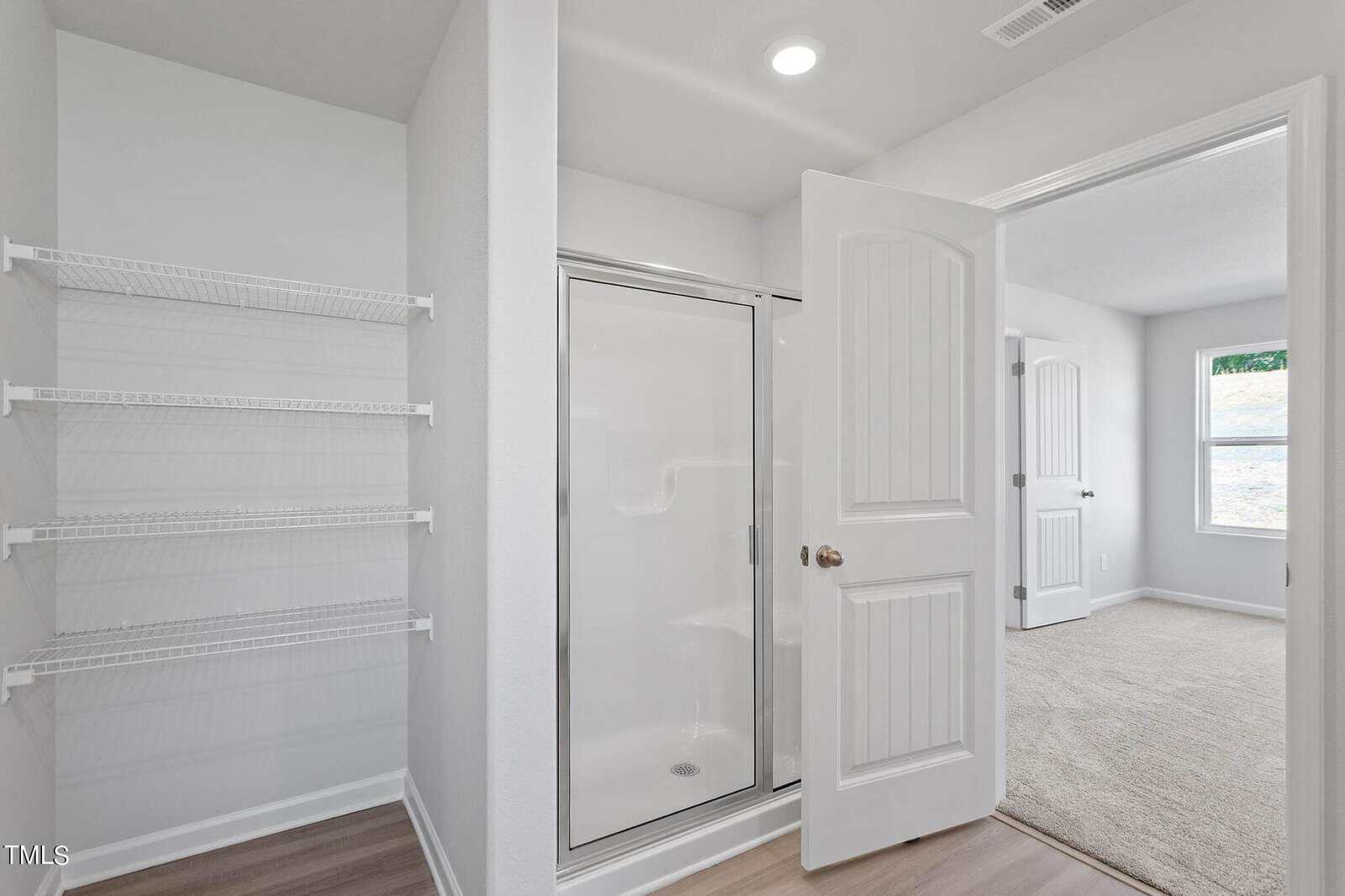 A minimalist bathroom with white walls features a glass-enclosed shower next to open wire shelving. The door is open to a bright carpeted bedroom with two windows, allowing natural light in, illuminating the wooden floor.