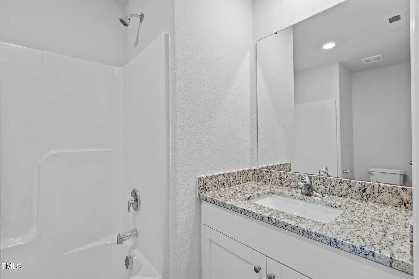 A modern bathroom featuring a white bathtub with a showerhead and a granite-topped vanity. The vanity has a white sink with a chrome faucet and a large mirror above it. The walls are painted white, creating a clean and bright atmosphere.