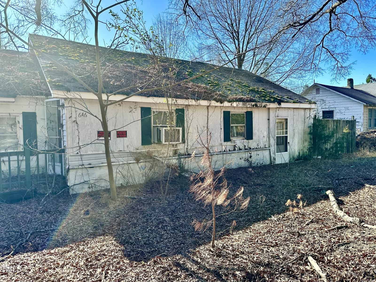 A weathered, single-story house with peeling white paint, an overgrown yard, and a window air conditioning unit, bathed in sunlight with shadows cast by bare branches.