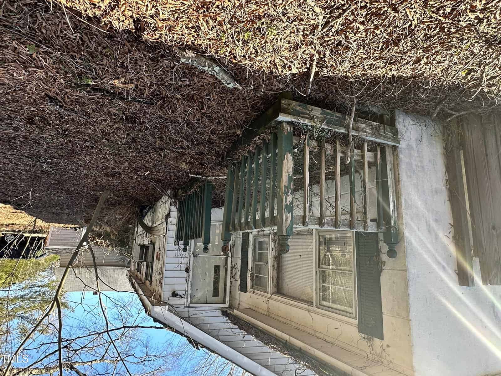 A house with white siding and green shutters is seen from an upside-down perspective with dry leaves and branches visible above, suggesting the photo was taken with the camera inverted.